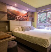 Un dormitorio moderno con una cama grande, una mesita de noche, un banco y una ventana con vistas a árboles.