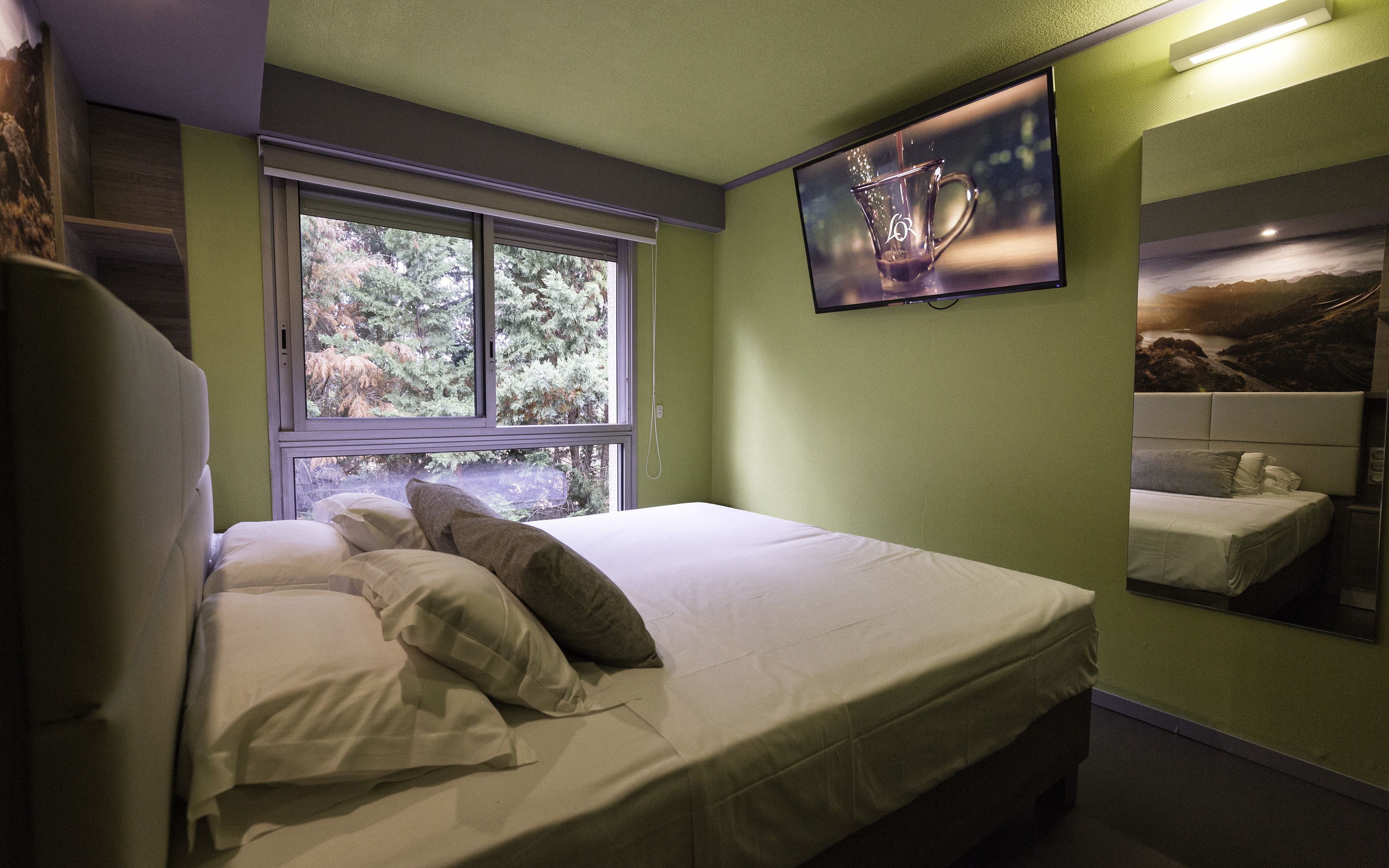Una cama bien hecha con almohadas, una ventana con vistas a árboles y un televisor fijado a la pared.