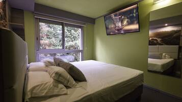 Una cama bien hecha con almohadas, una ventana con vistas a árboles y un televisor fijado a la pared.