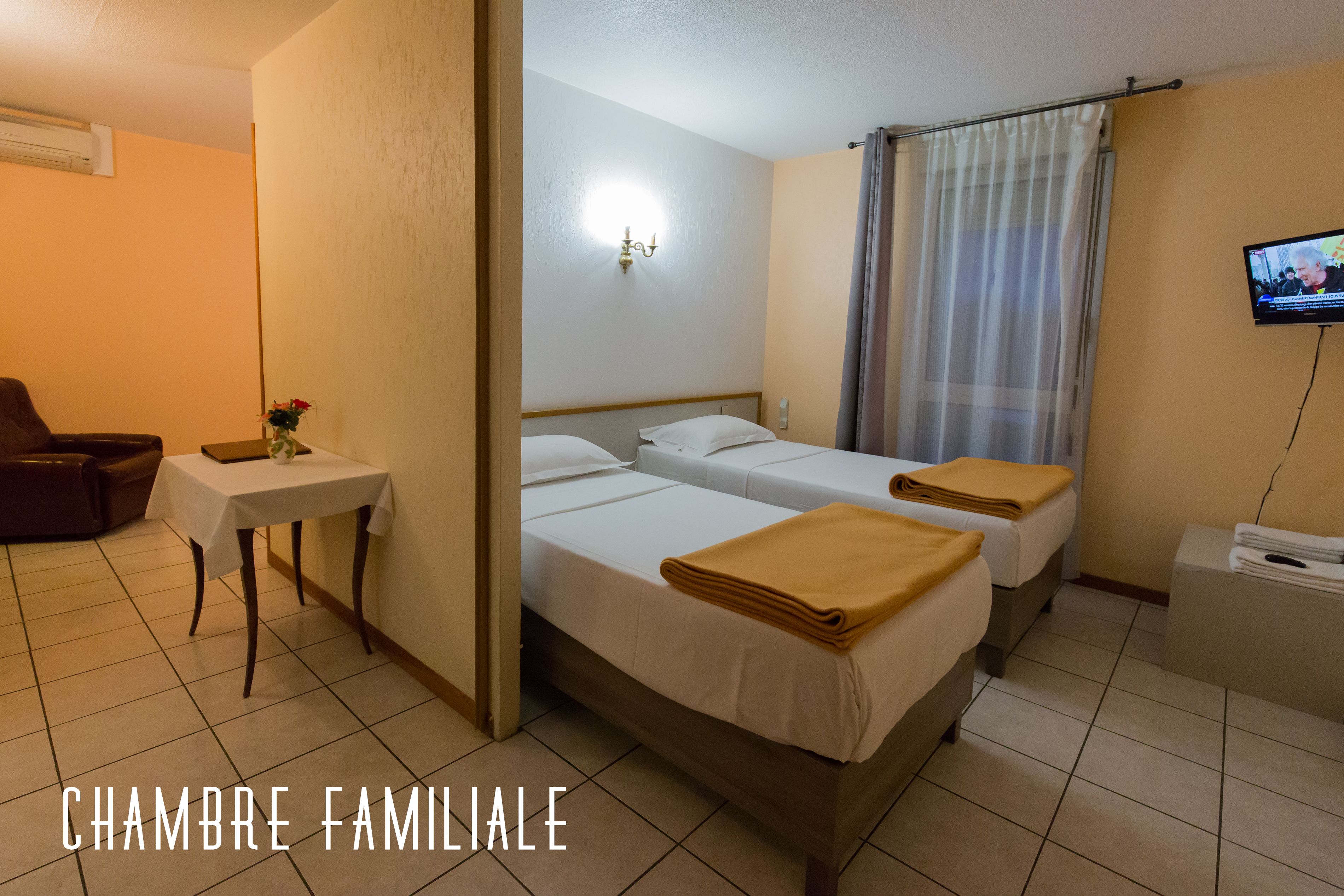 Ein Familienzimmer mit zwei Betten, einem kleinen Tisch mit einer Vase, einem Sessel und einem Fernseher an der Wand.