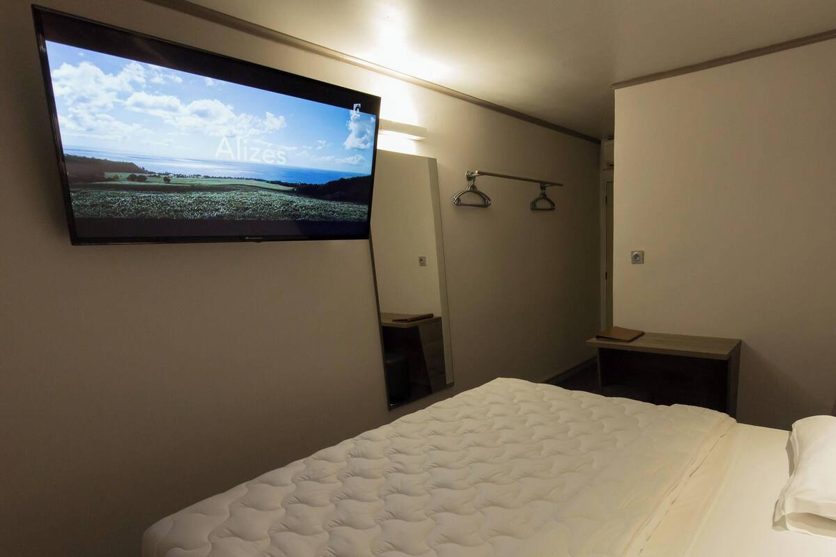 A hotel room with a bed, a TV mounted on the wall, a mirror, and a wooden shelf.