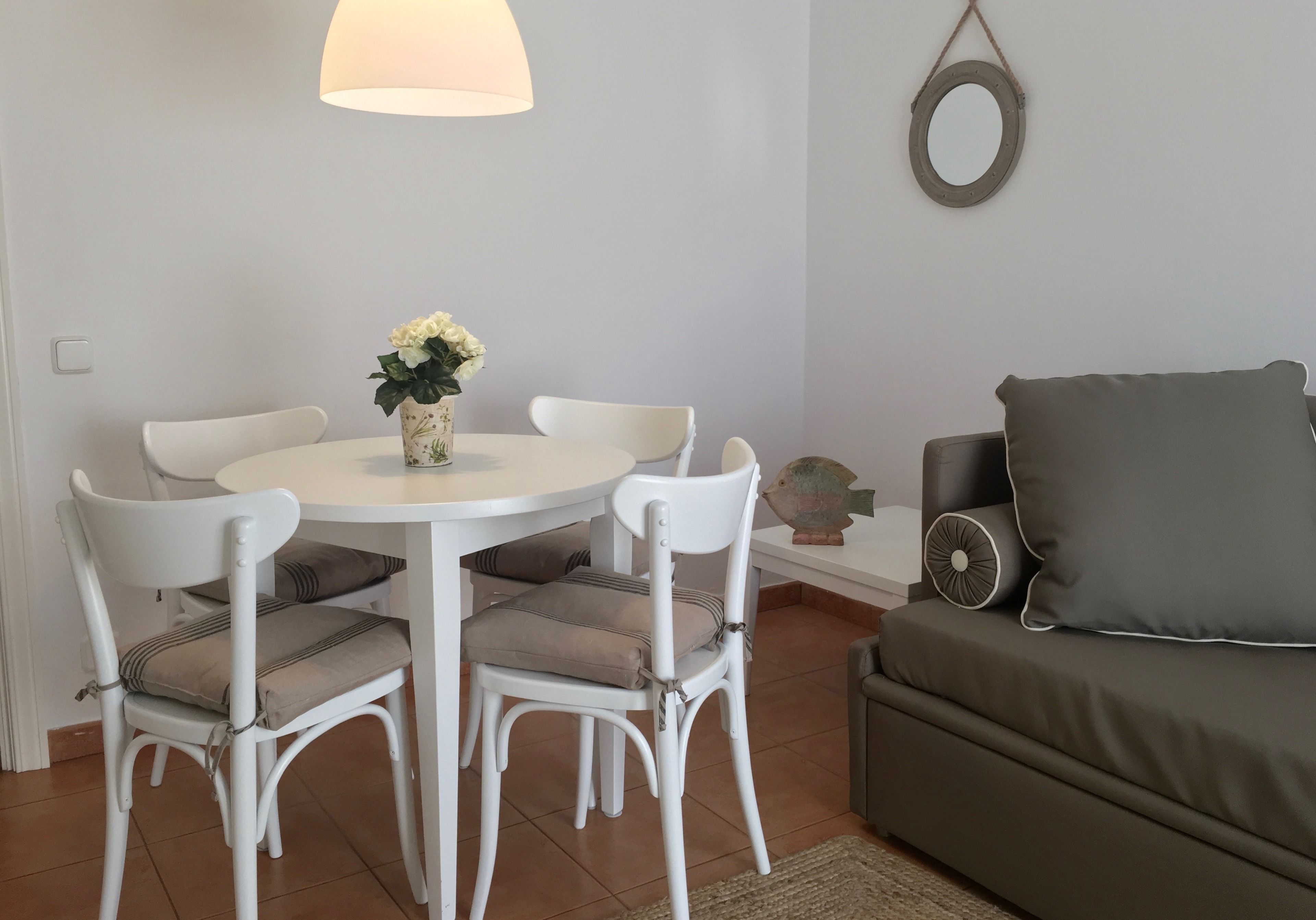 Un coin repas avec une table ronde, des chaises blanches et un vase rempli de fleurs. On y trouve également un canapé gris et un petit objet décoratif accroché au mur.