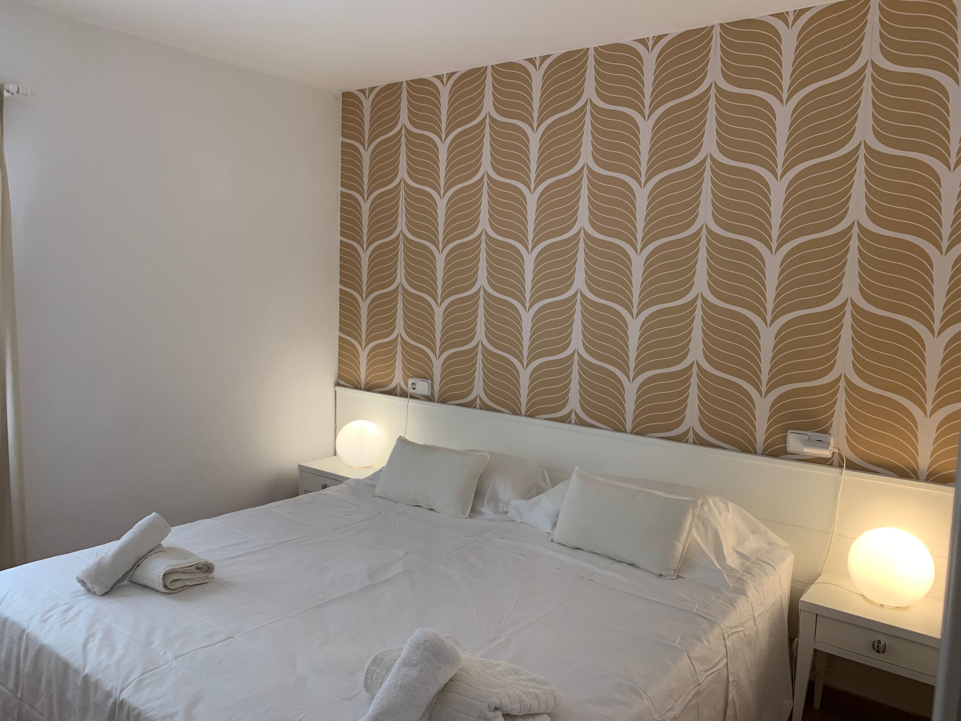A neatly made bed with white linens and pillows, a wall with a leaf-pattern wallpaper, and bedside lamps.