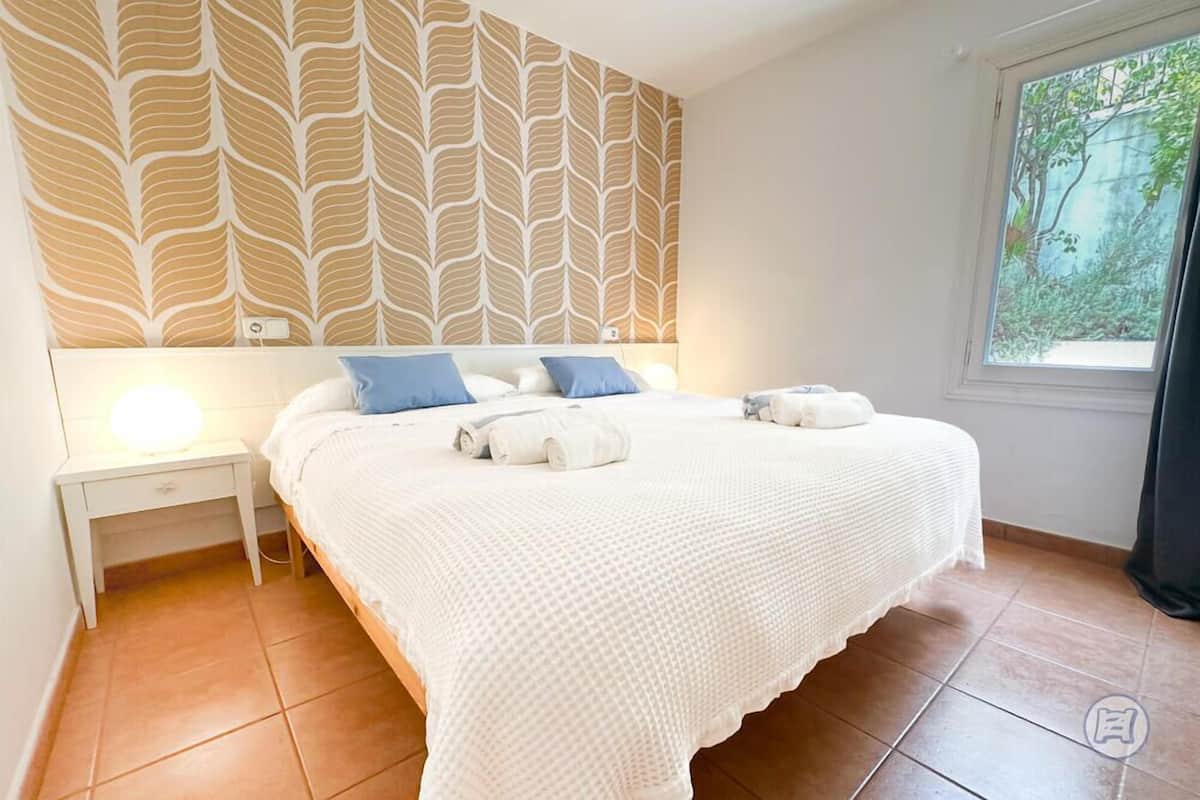 Un dormitorio con una cama grande, ropa de cama blanca, una mesita de noche, un papel tapiz estampado y una ventana con vistas a zonas verdes.