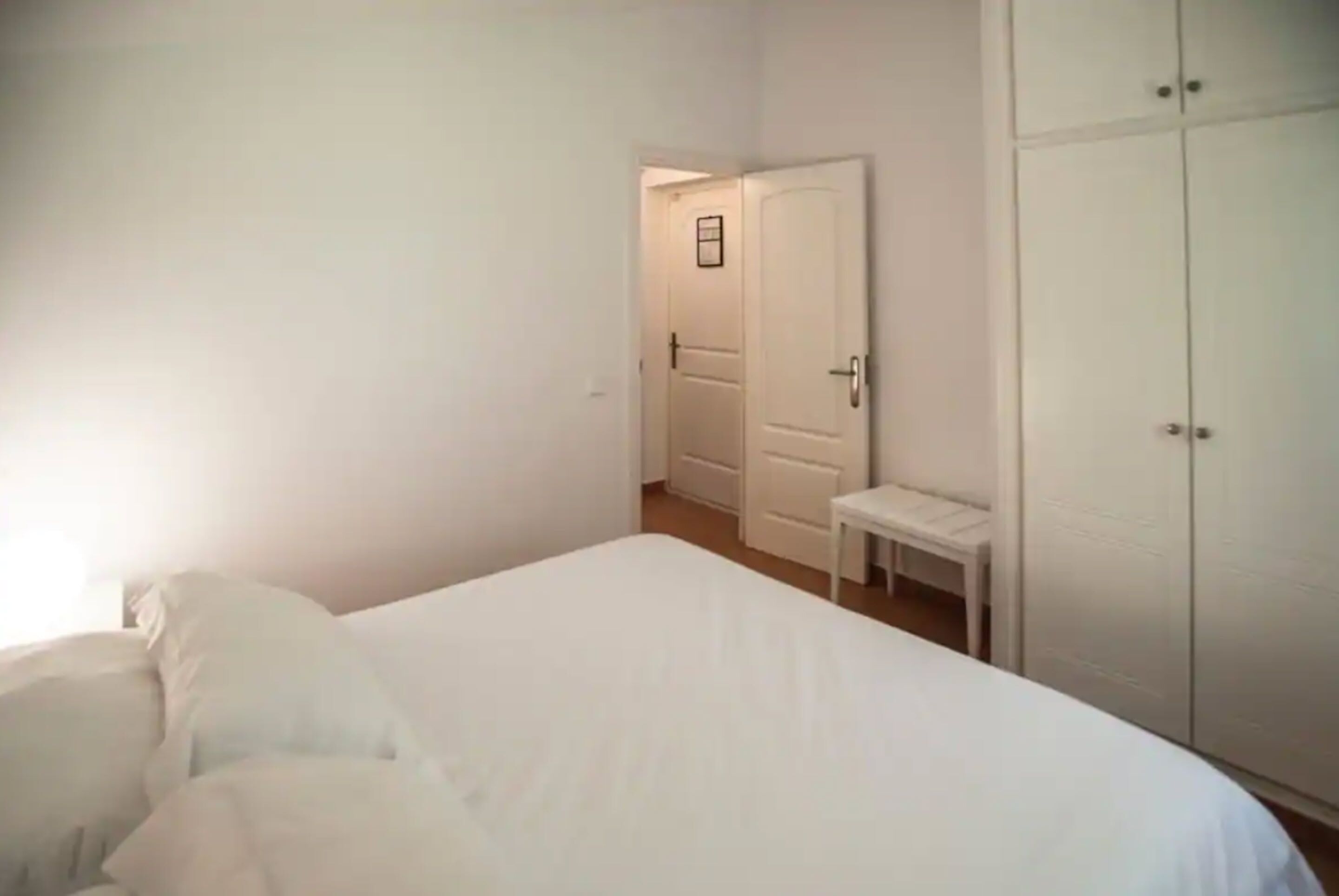A bedroom with a bed, a bedside table, a wardrobe, and a door leading to another room.
