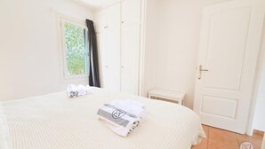 Una cama bien hecha con una toalla doblada y una colcha, una ventana con cortinas y una puerta cerrada.