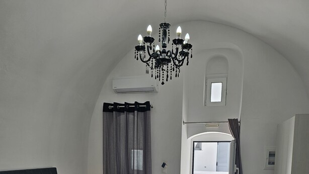 A bedroom with a bed, a chandelier, a window with curtains, and a view of an outdoor area. - Unique Leta (Santorini)