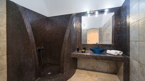 A modern bathroom with a curved shower area, a sink with a blue bowl, and a mirror above the sink. - Unique Leta (Santorini)