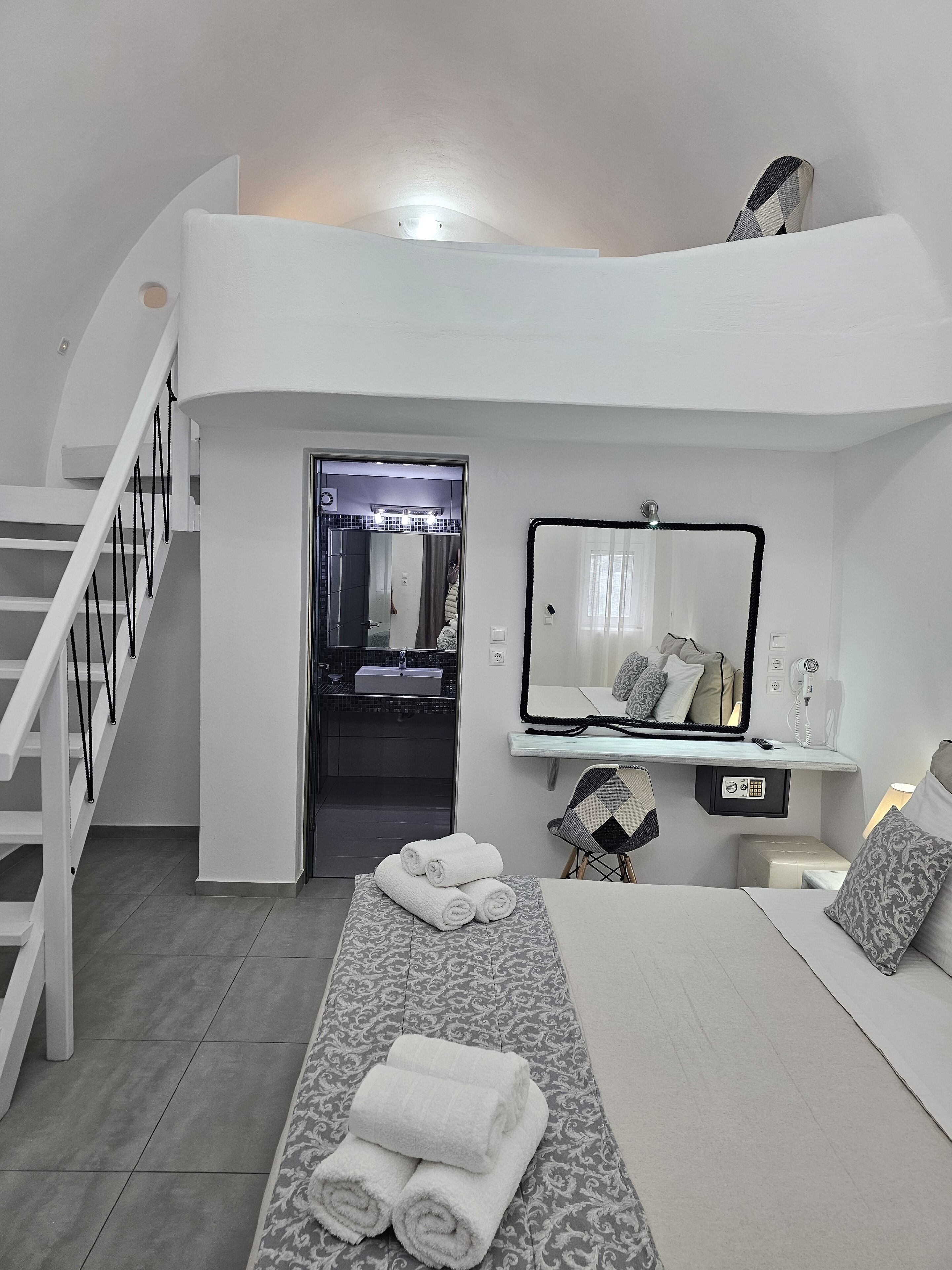 Una habitación de hotel moderna con una cama, una mesita de noche, un espejo, un escritorio pequeño con una silla, un baño con lavamanos y espejo, y una escalera.