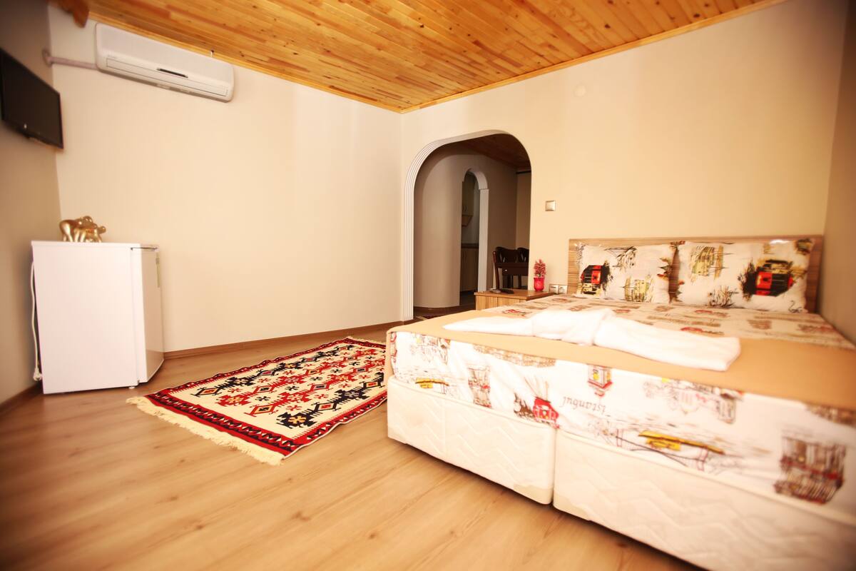 A bedroom with a wooden ceiling, a bed with a patterned headboard, a white refrigerator, and a red and black rug.