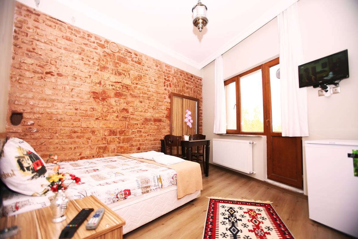 A room with a brick wall, a bed with a patterned bedspread, a wooden desk, a chair, a small table with a vase and remote controls, and a television on the wall.