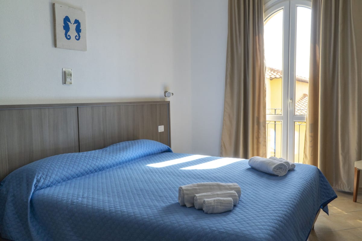 A hotel room with a large bed, a headboard, a wall-mounted thermostat, and a window with curtains.