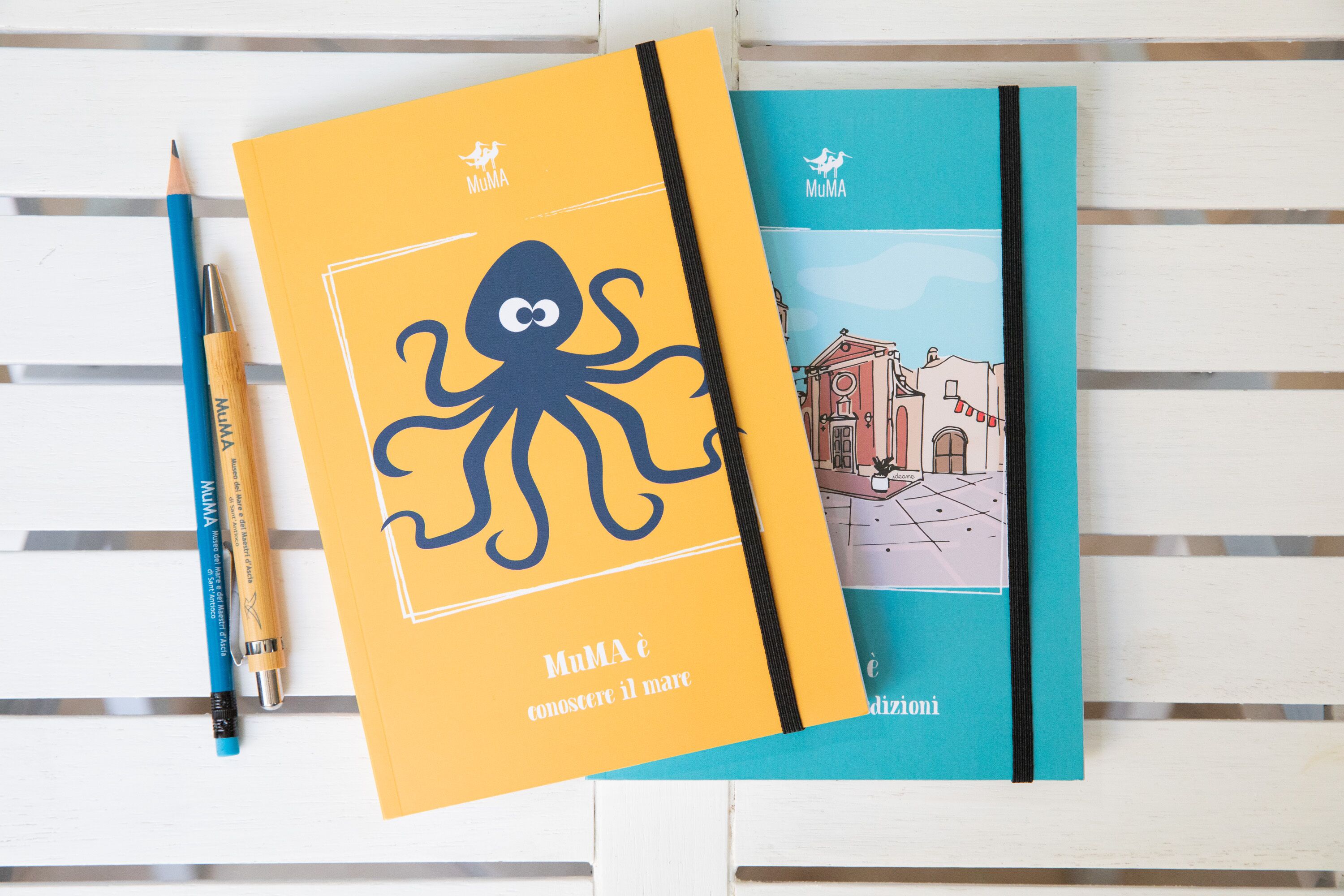 Two notebooks with illustrations, one with an octopus and the other with a cityscape, and a blue pencil on a white wooden surface.