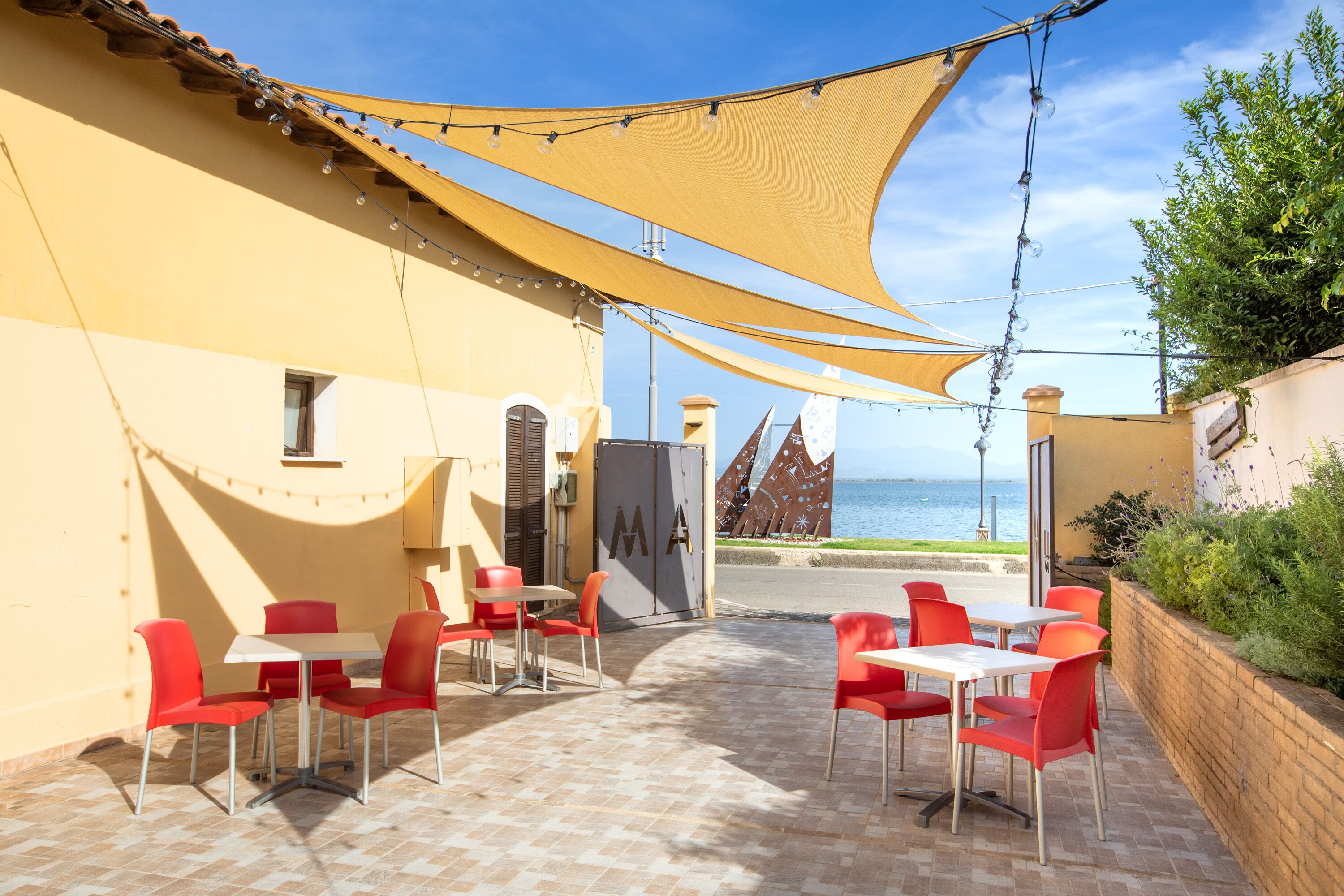 Un patio al aire libre con sillas y mesas rojas, un edificio amarillo y vistas al mar.