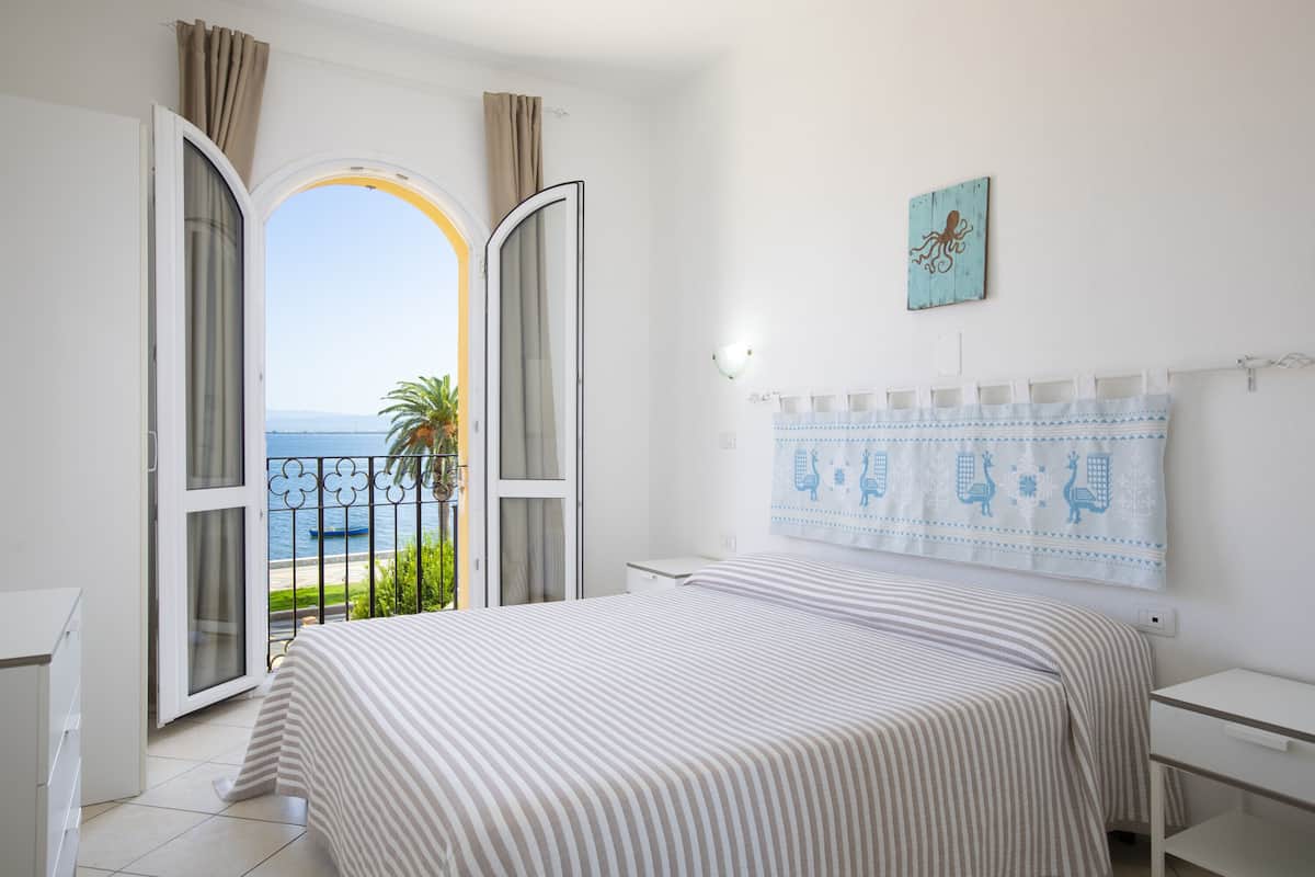 A bedroom with a bed, a nightstand, a window with curtains, a wall-mounted light, and a view of the sea.