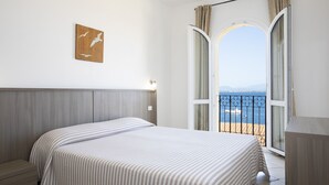 Een slaapkamer met een bed, een nachtkastje, uitzicht op zee door een balkondeur en een kunstwerk aan de muur.