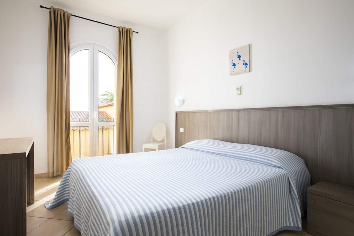 A bedroom with a bed, a chair, a desk, a window with curtains, and a wall-mounted light fixture.