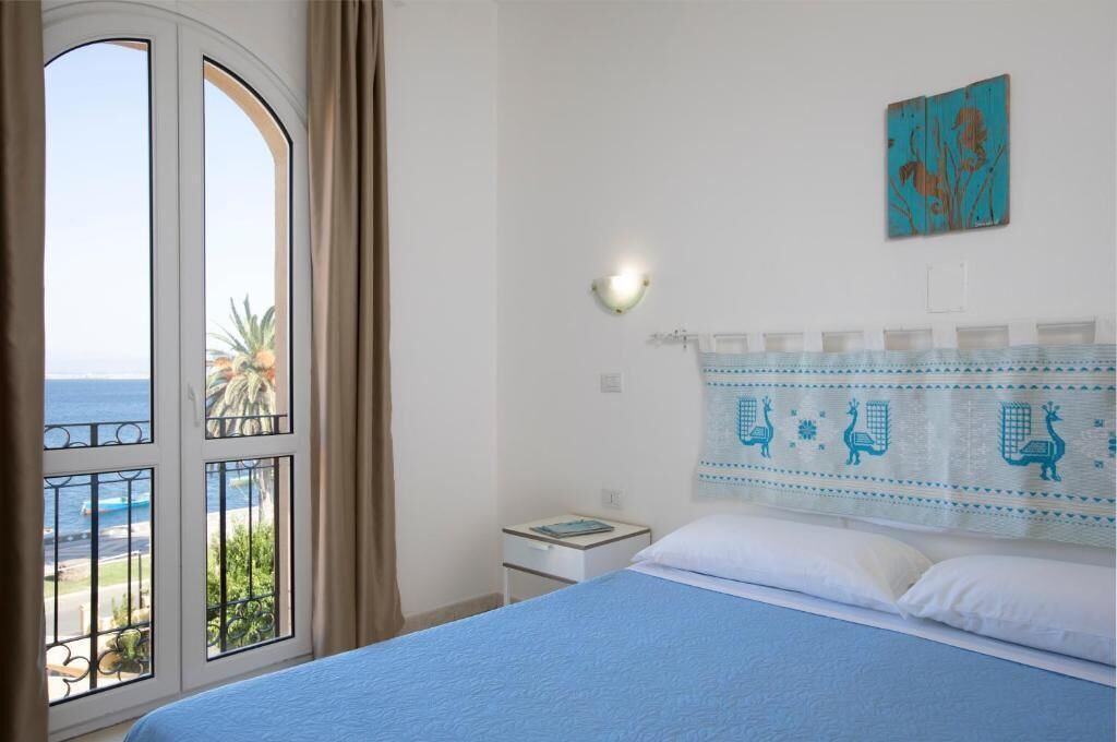 A bedroom with a bed, a nightstand, a wall-mounted lamp, a window with curtains, and a view of the sea.