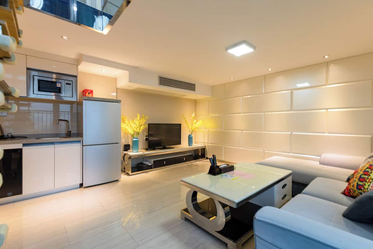 A modern kitchen with a microwave, refrigerator, and built-in oven. There is a dining area with a table and chairs, and a living room with a sofa and a TV.