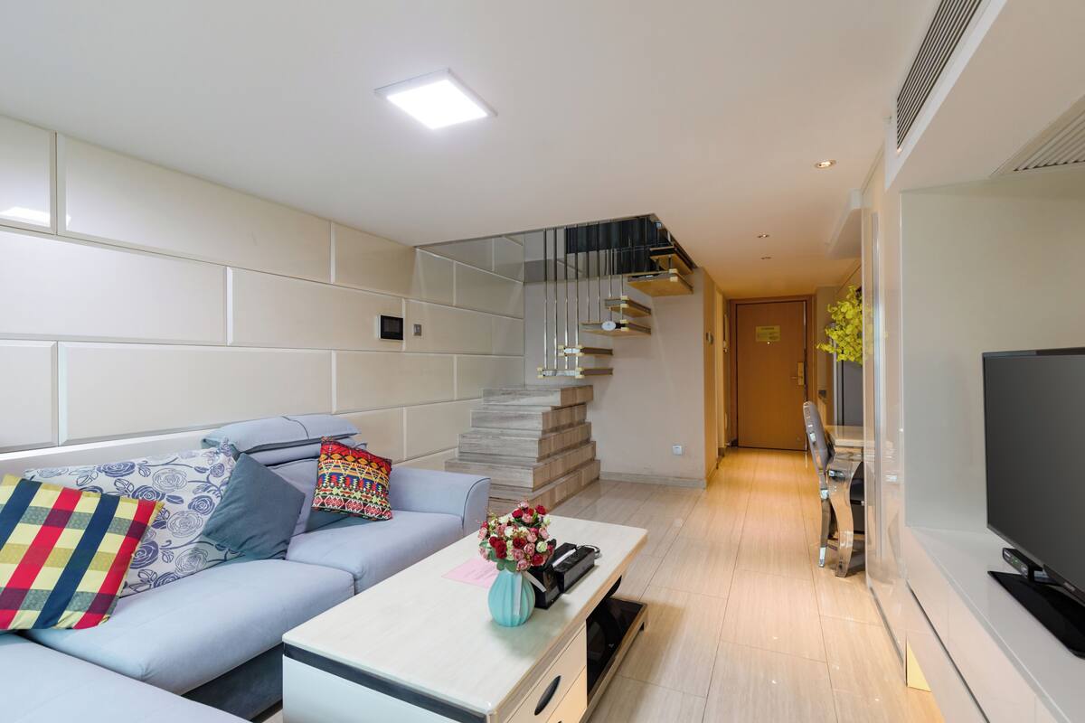 A modern living room with a light blue sofa, a white coffee table with a vase of flowers, a staircase with a glass railing, and a flat-screen TV on the right.