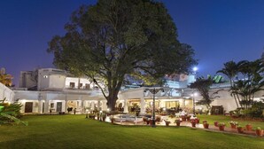 A large tree in front of a building with multiple balconies and a well-lit outdoor area with seating and potted plants. - Jehan Numa Palace Hotel (Bhopal)