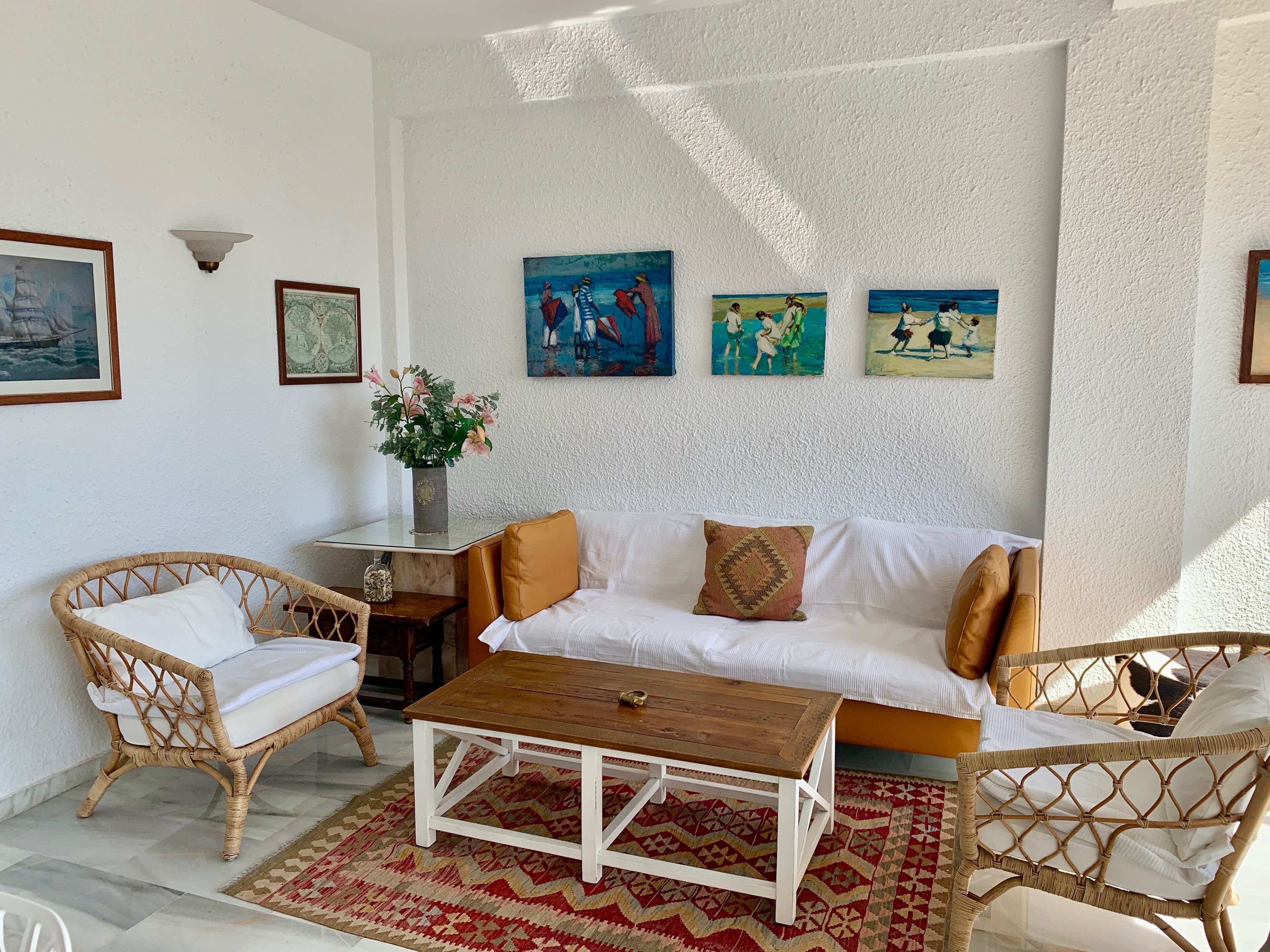 Ein Wohnzimmer mit einem weißen Sofa, zwei Korbstühlen, einem Holztisch und einem gemusterten Teppich. An den Wänden hängen Bilder und ein Blumenarrangement steht in einer Vase.