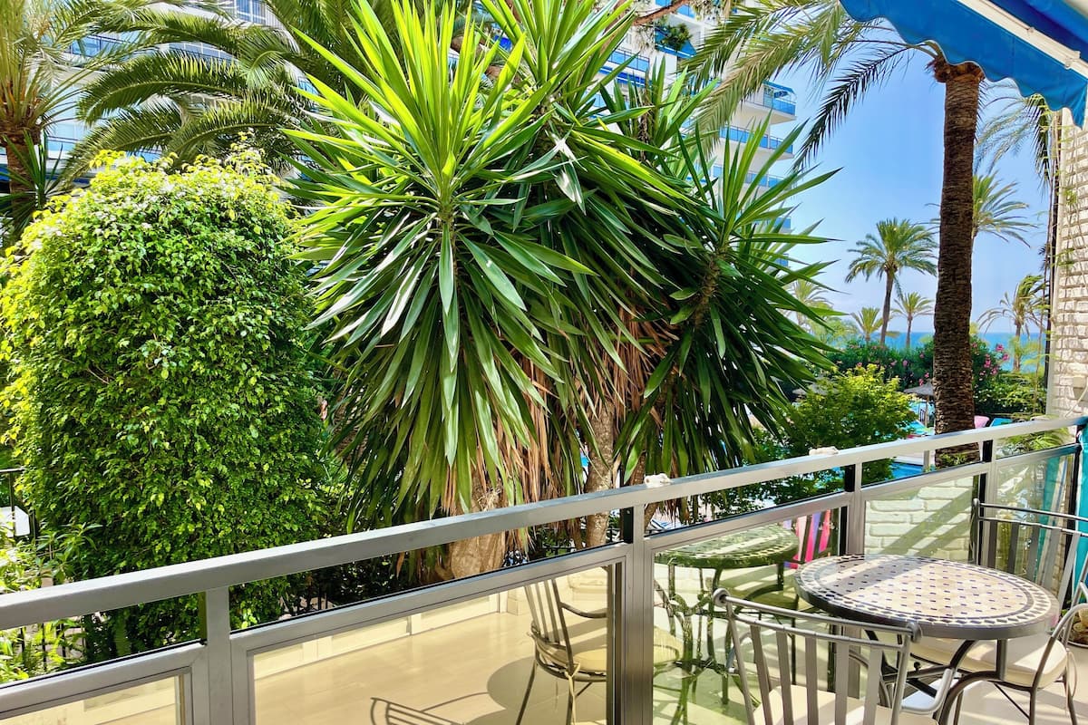 A balcony with a glass railing, a round metal table, and chairs, overlooking a lush garden with various plants and a building in the background.