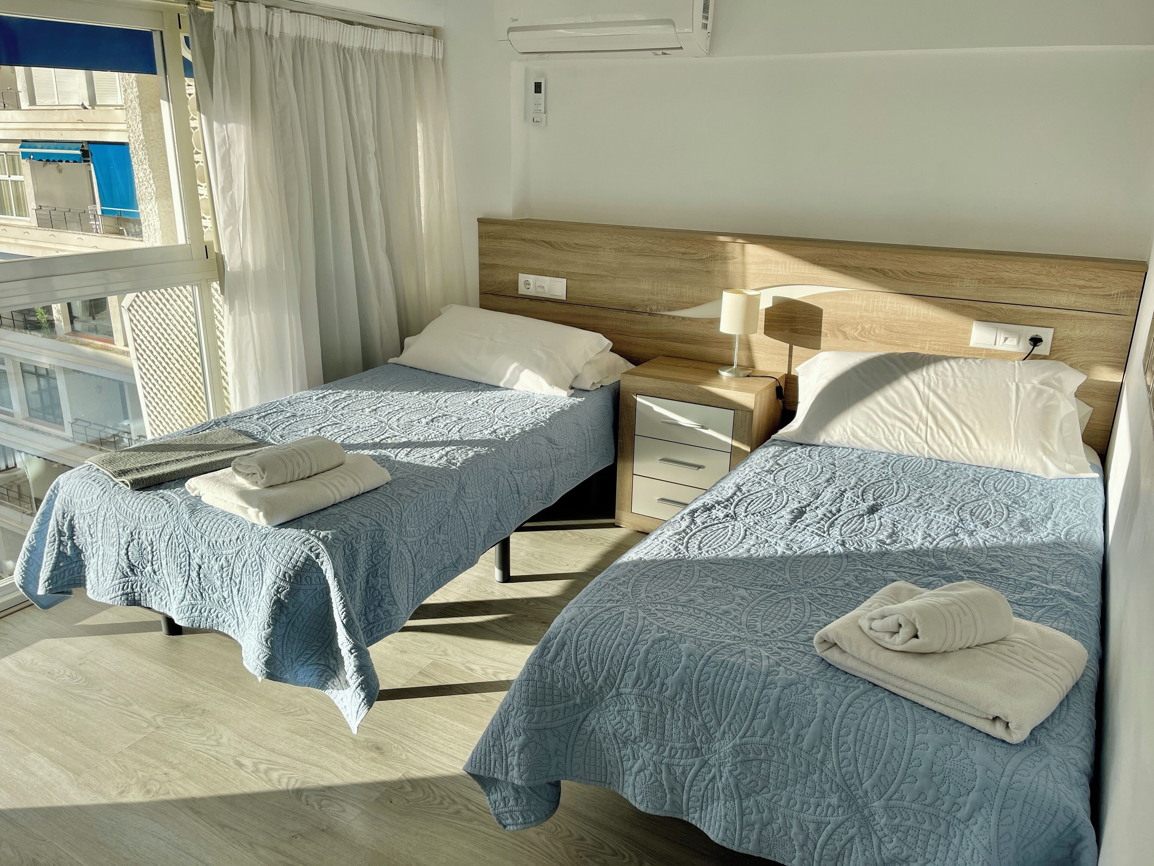 Une chambre d’hôtel avec deux lits, chacun recouvert d’une courtepointe matelassée bleue et agrémenté d’oreillers blancs. On y trouve une tête de lit en bois, une table de chevet avec une lampe et un petit tiroir. La chambre est dotée d’une fenêtre avec des rideaux et offre une vue sur un bâtiment à l’extérieur.