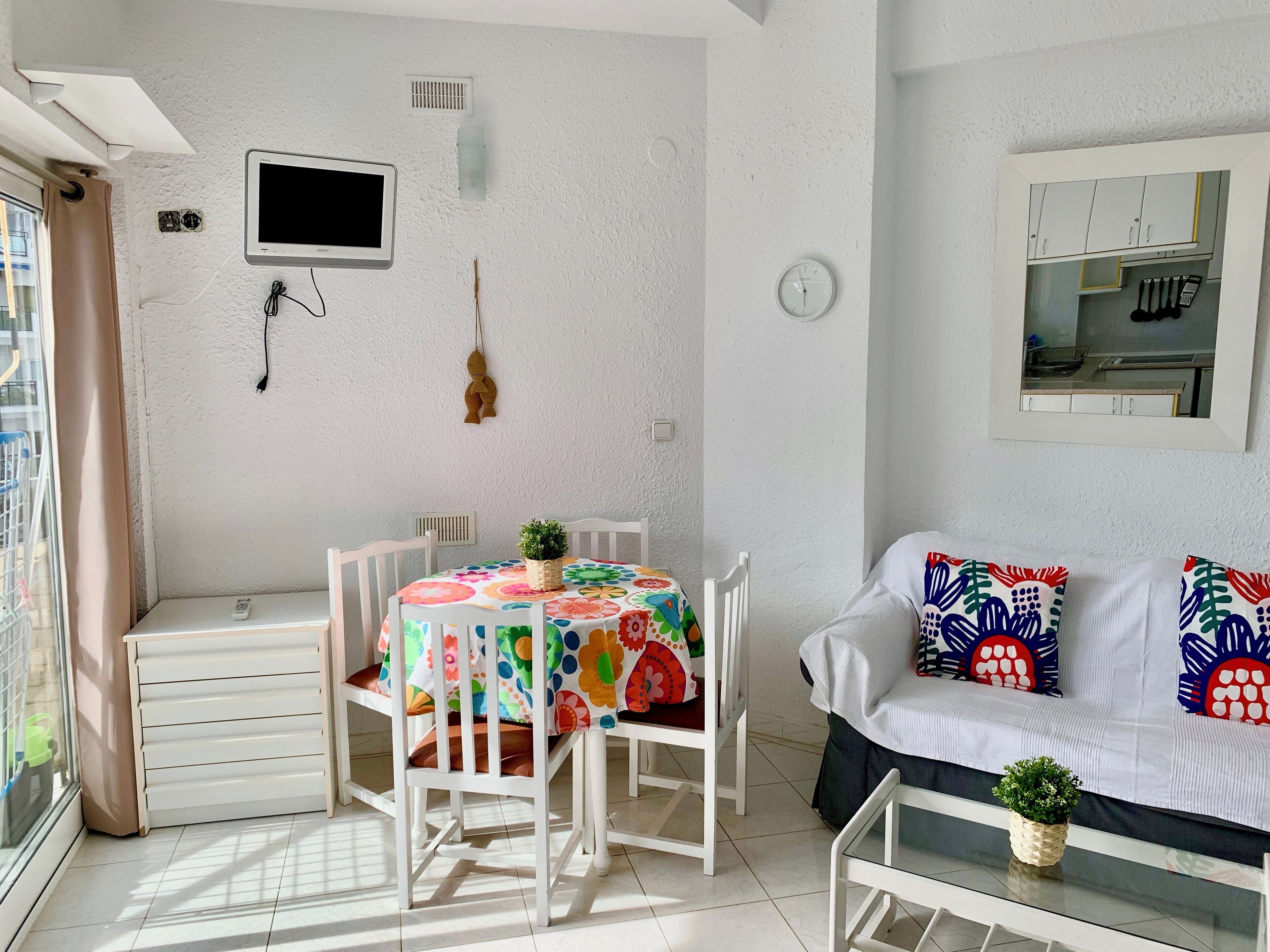 A small, modern room with a dining table and chairs, a sofa, a TV, and a kitchen area visible through a glass door.