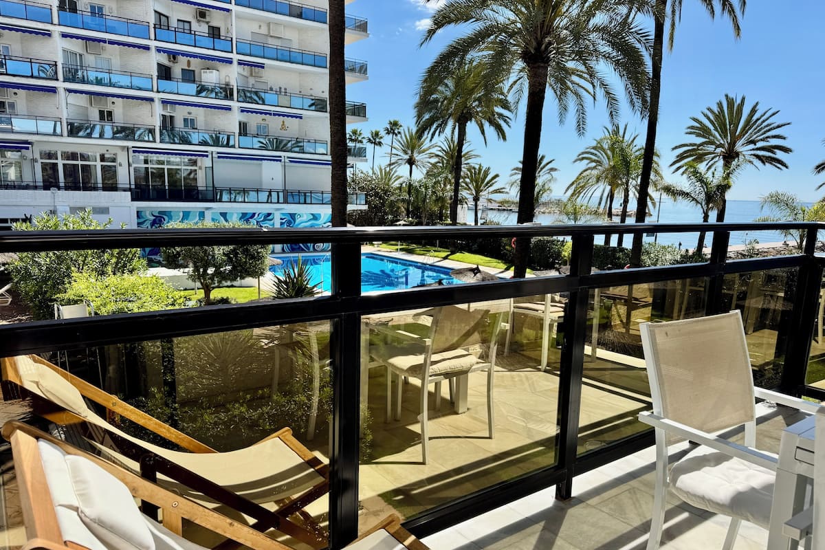 A balcony with a view of a swimming pool, palm trees, and a modern building in the background.
