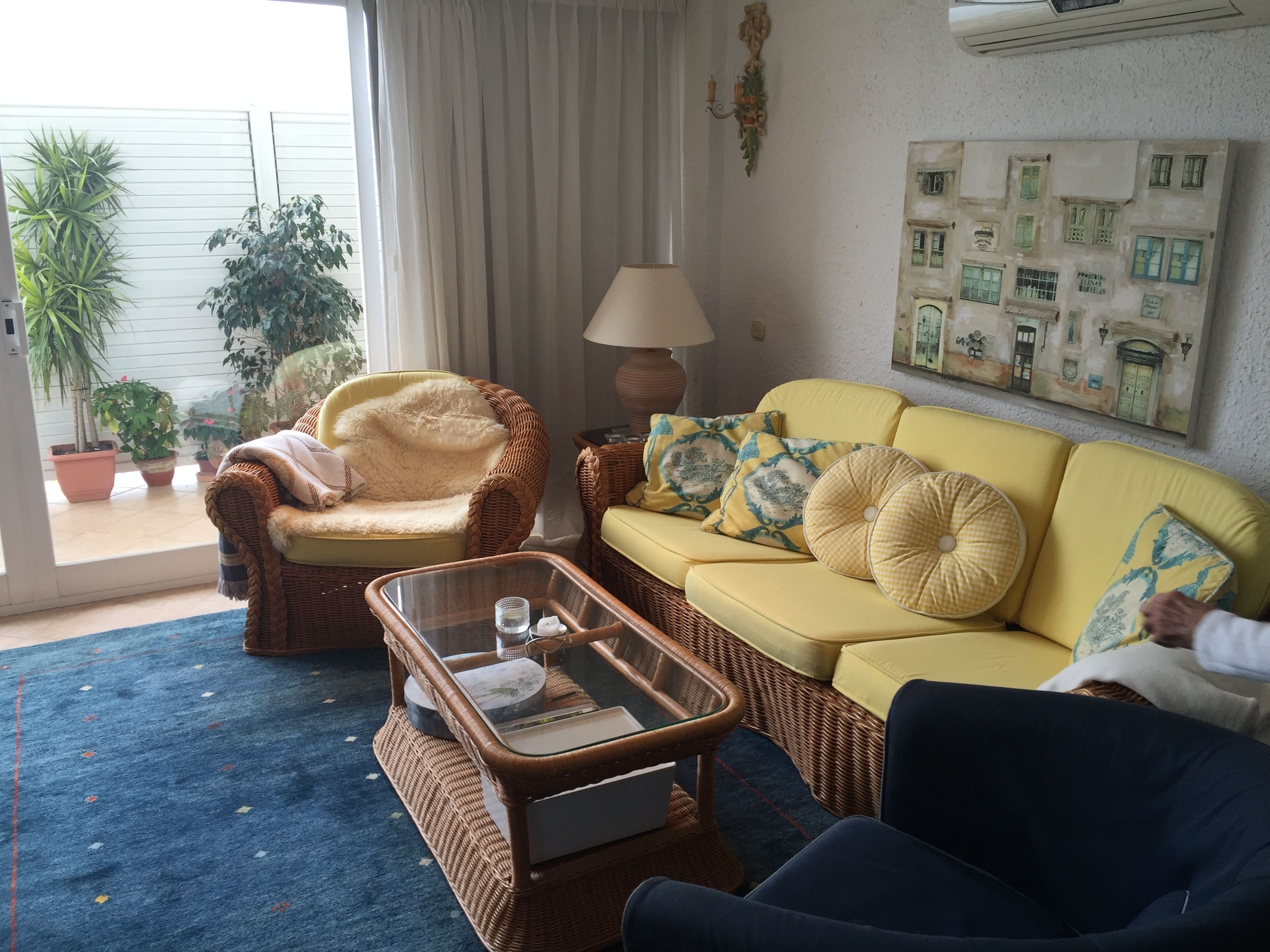 A living room with a yellow sofa, a wicker armchair, and a glass coffee table. There are potted plants and a wall art piece.