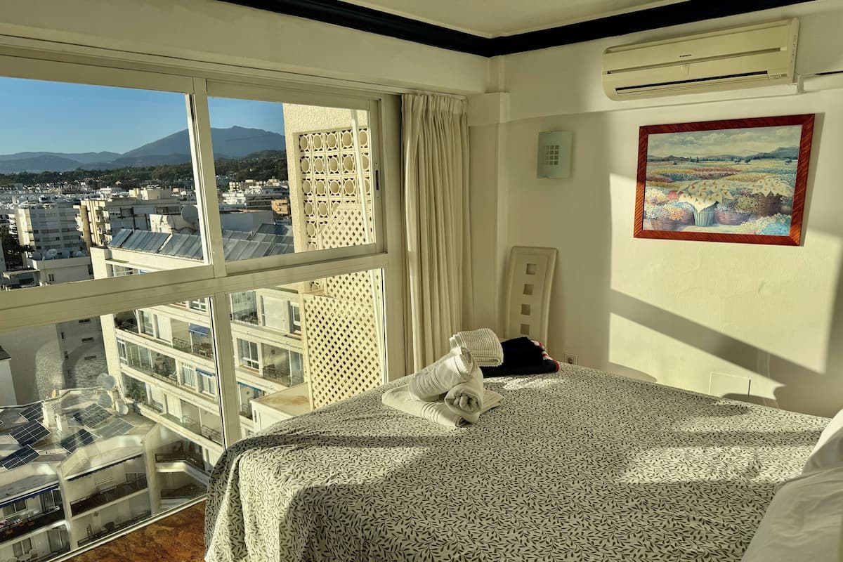 A hotel room with a large window, a bed with a patterned bedspread, a painting on the wall, and an air conditioning unit.