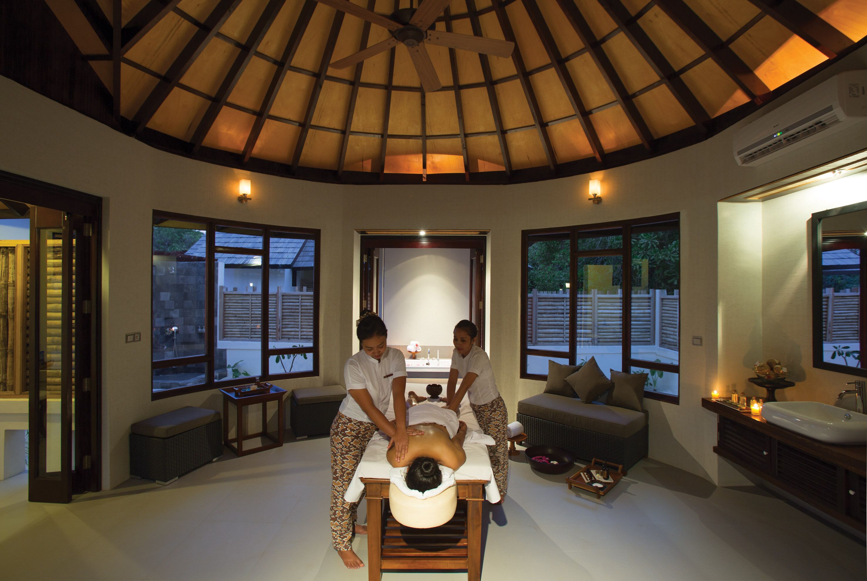 Sala de massagem com teto de madeira, janelas amplas, mesa de massagem e um sofá.