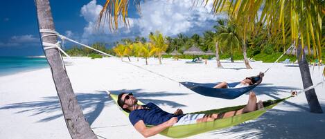 A beach scene with two hammocks tied between palm trees, white sand, and clear blue water.