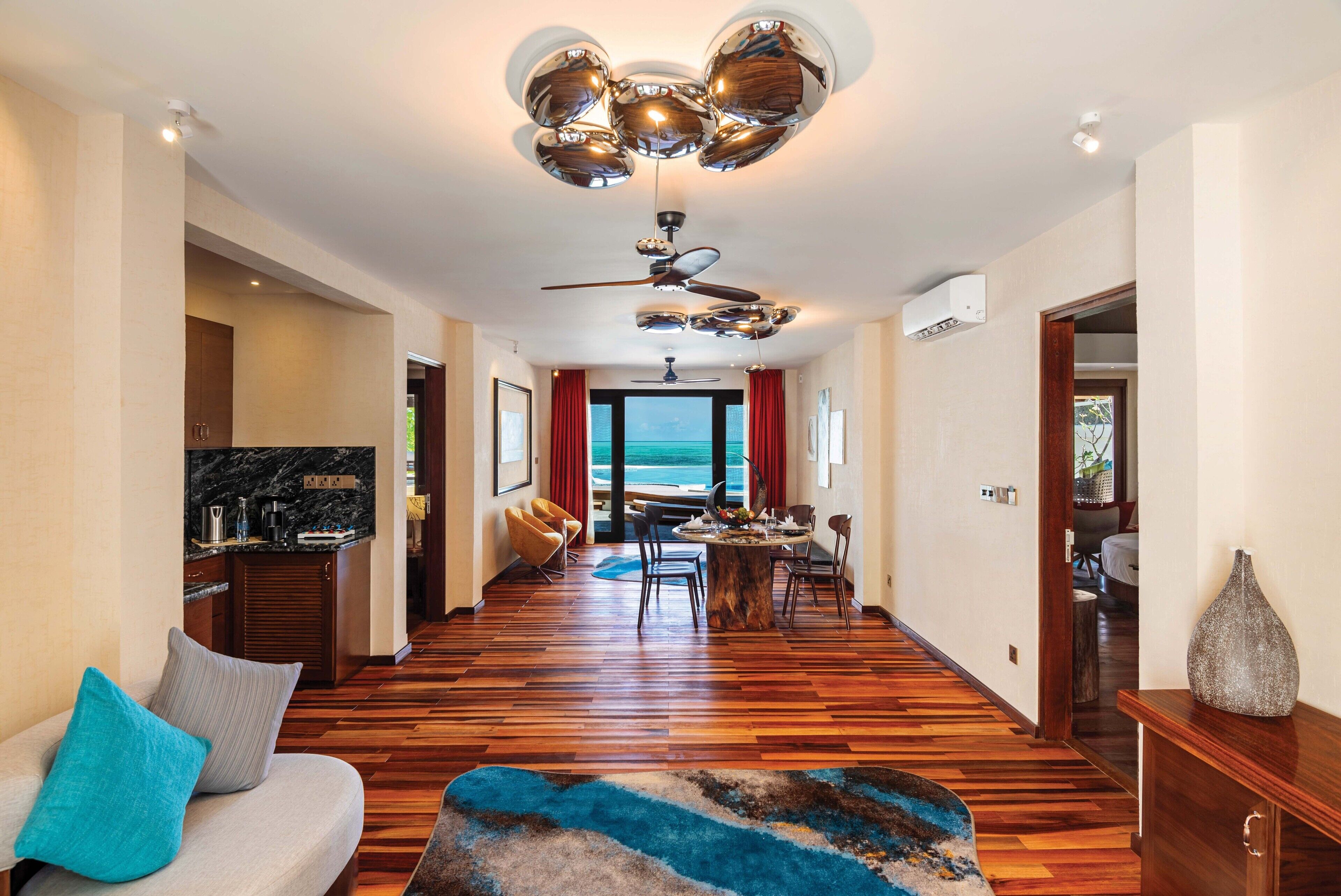 A spacious living area with a wooden floor, a sofa with a blue pillow, a dining table with chairs, and a ceiling fan.