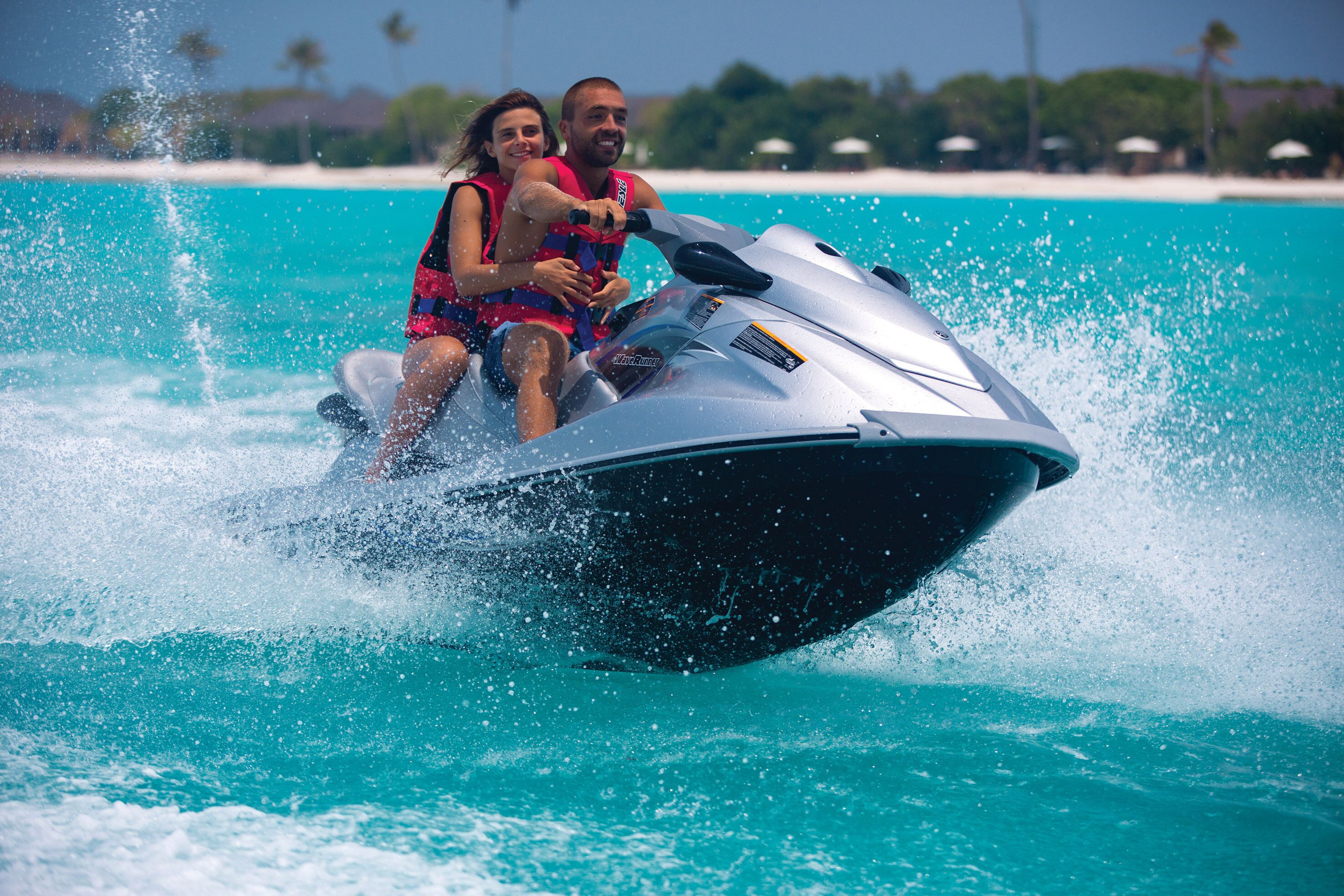 Une personne conduisant un jet ski sur une eau turquoise, avec une plage et des palmiers en arrière-plan.