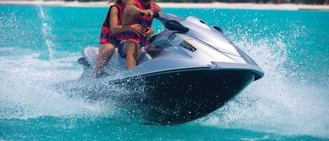 Une personne conduisant un jet ski sur une eau turquoise, avec une plage et des palmiers en arrière-plan.