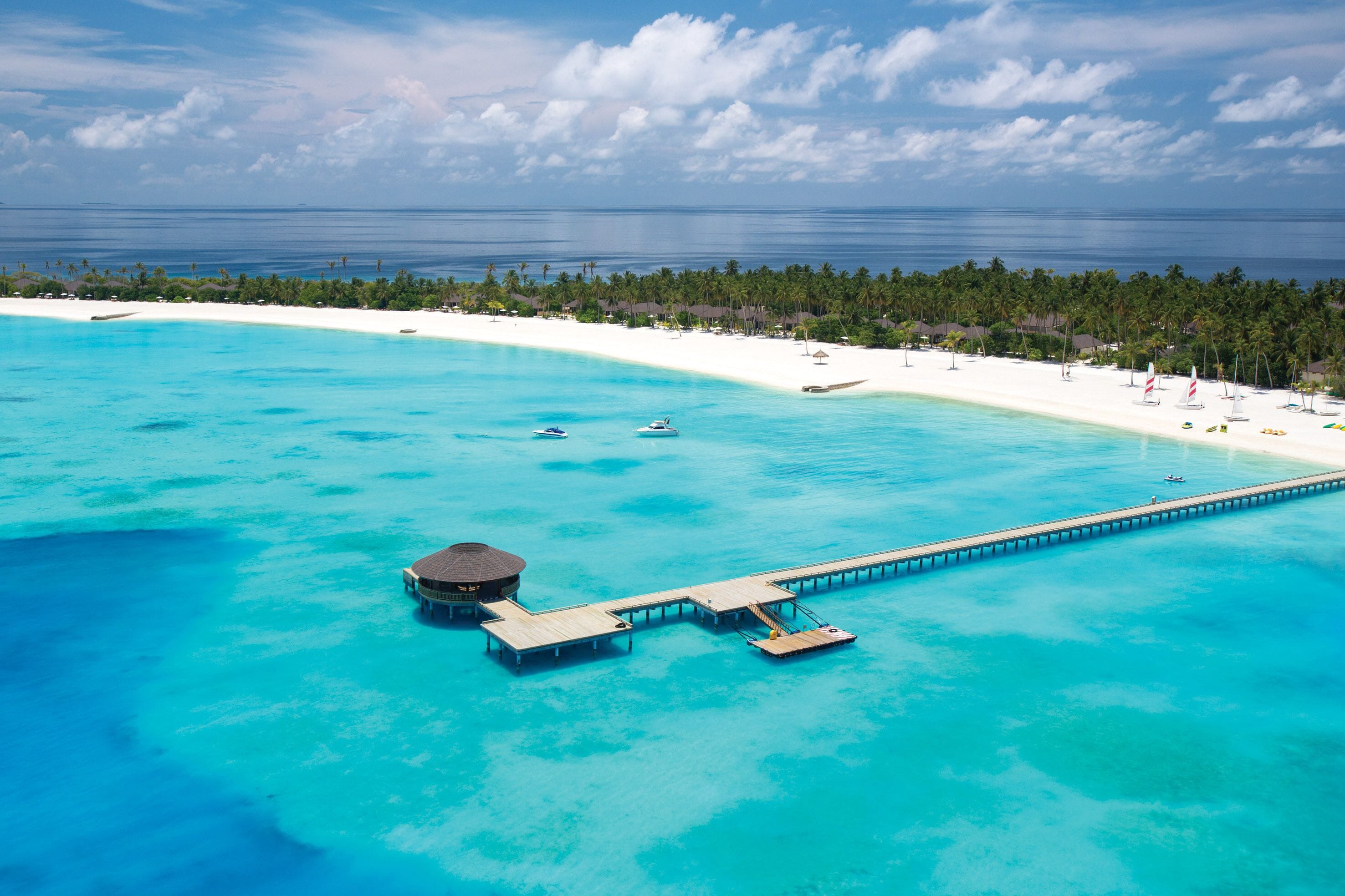 Vue aérienne d’un complexe hôtelier en bord de mer, avec une plage de sable blanc, des eaux turquoise et une longue jetée menant à une petite structure construite sur pilotis.