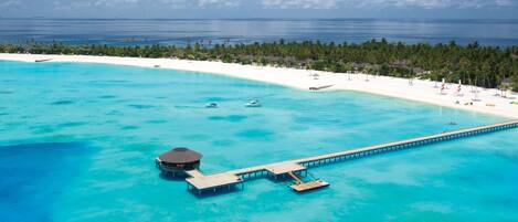 Vue aérienne d’un complexe hôtelier en bord de mer, avec une plage de sable blanc, des eaux turquoise et une longue jetée menant à une petite structure construite sur pilotis.