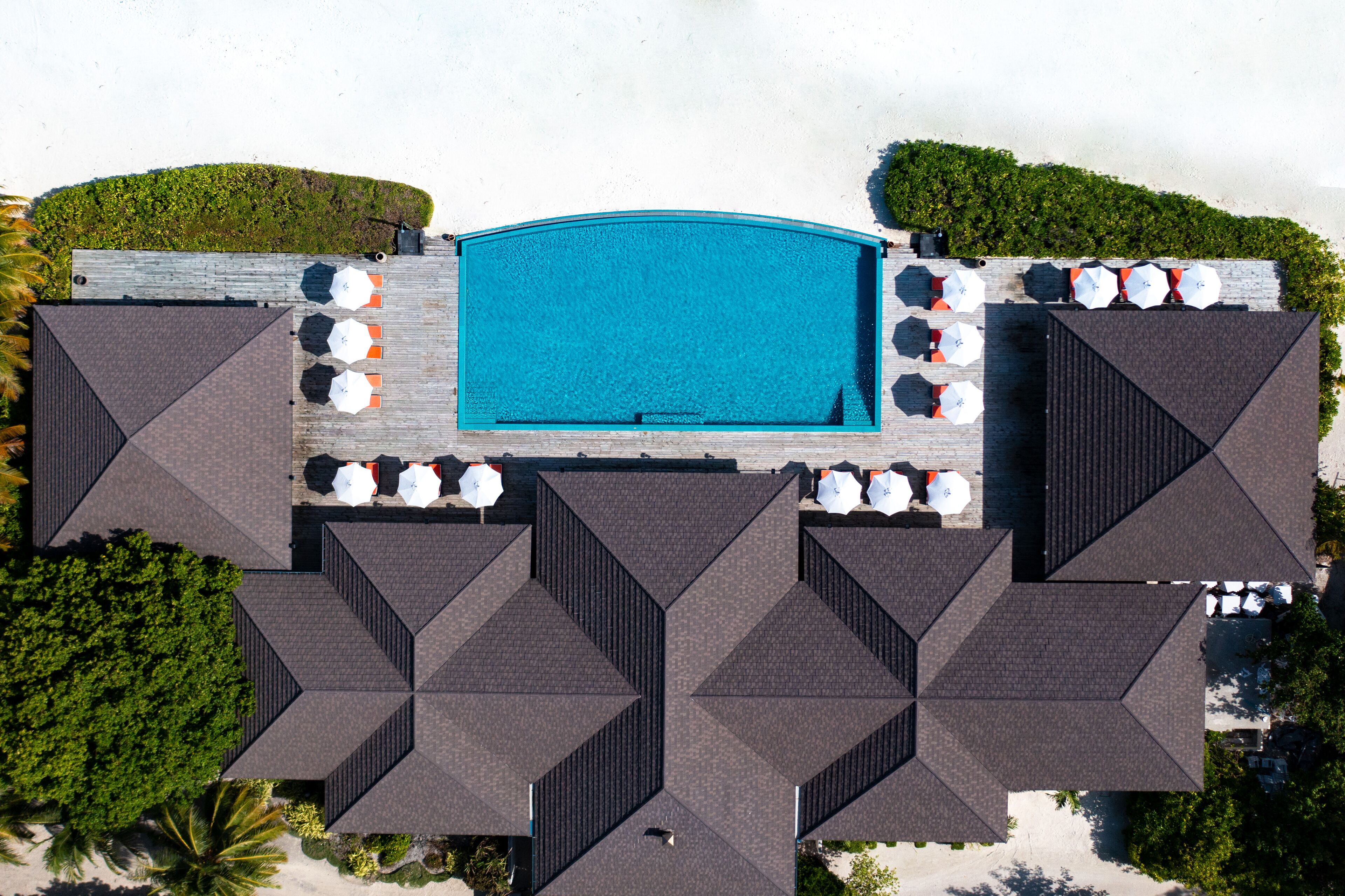 An aerial view of a hotel with a central swimming pool, multiple outdoor seating areas with umbrellas, and a symmetrical roof design.