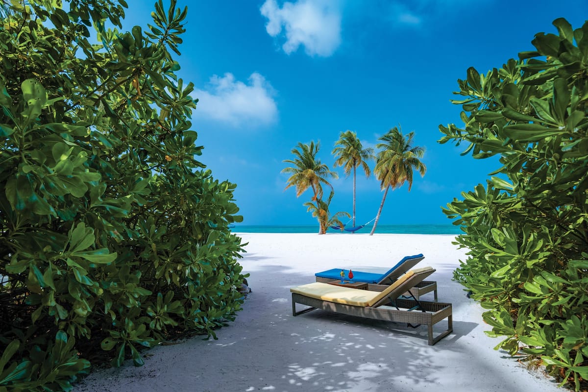 Eine Strandansicht mit Liegestuhl und Hängematte, Palmen und einem strahlend blauen Himmel.
