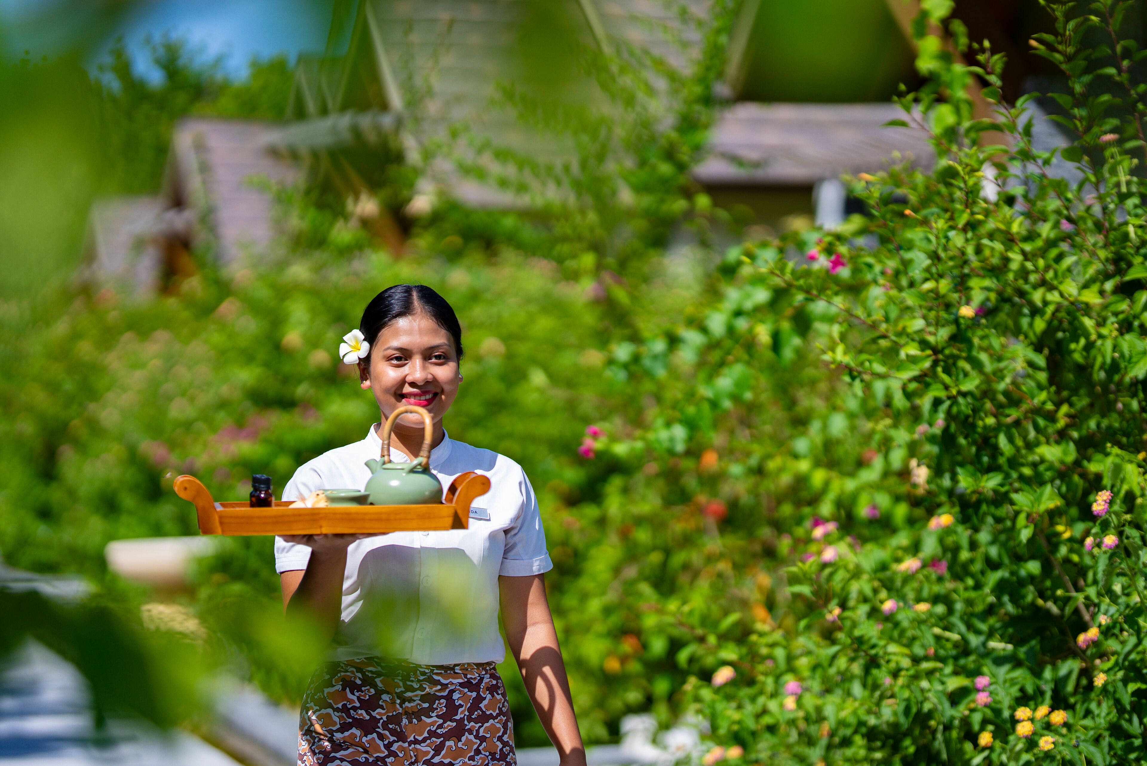 A person carrying a tray with a teapot and cups, surrounded by lush greenery and a traditional house in the background.