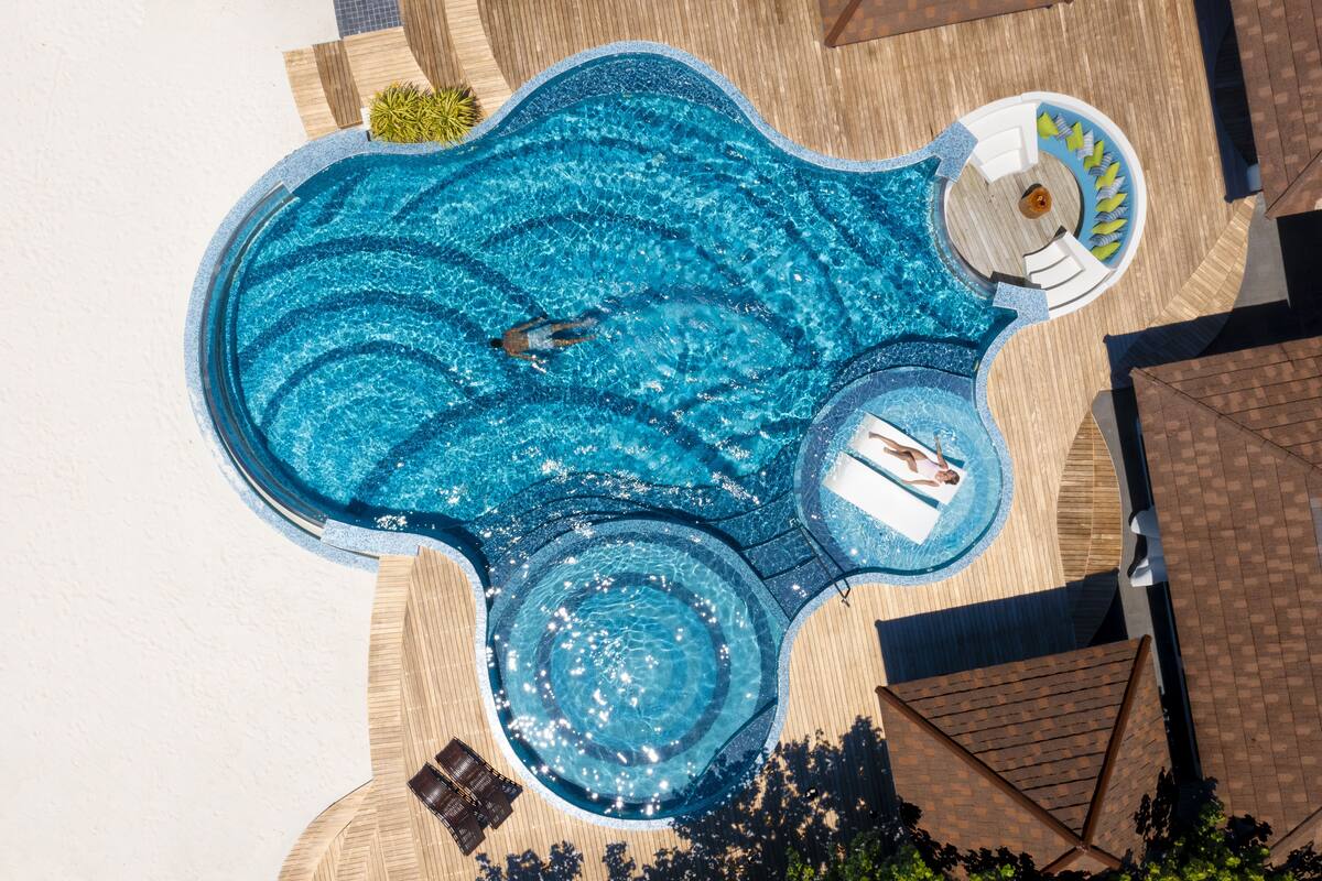 Ein großer, ungewöhnlich geformter Pool mit verschiedenen Bereichen, darunter ein Whirlpool, umgeben von einer Holzterrasse und Liegestühlen.