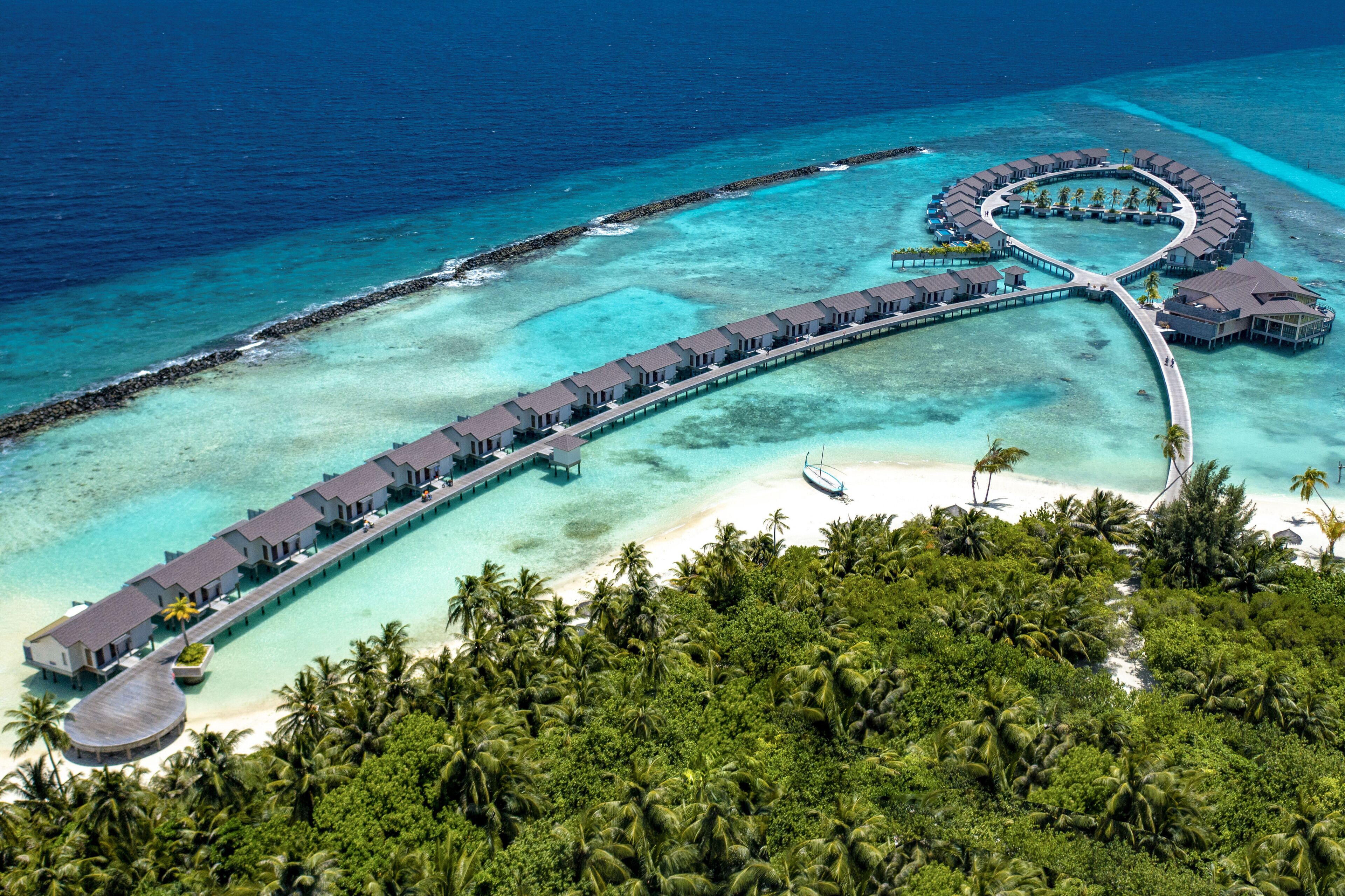 An overwater resort with a crescent-shaped building, connected by walkways, surrounded by turquoise waters and palm trees.