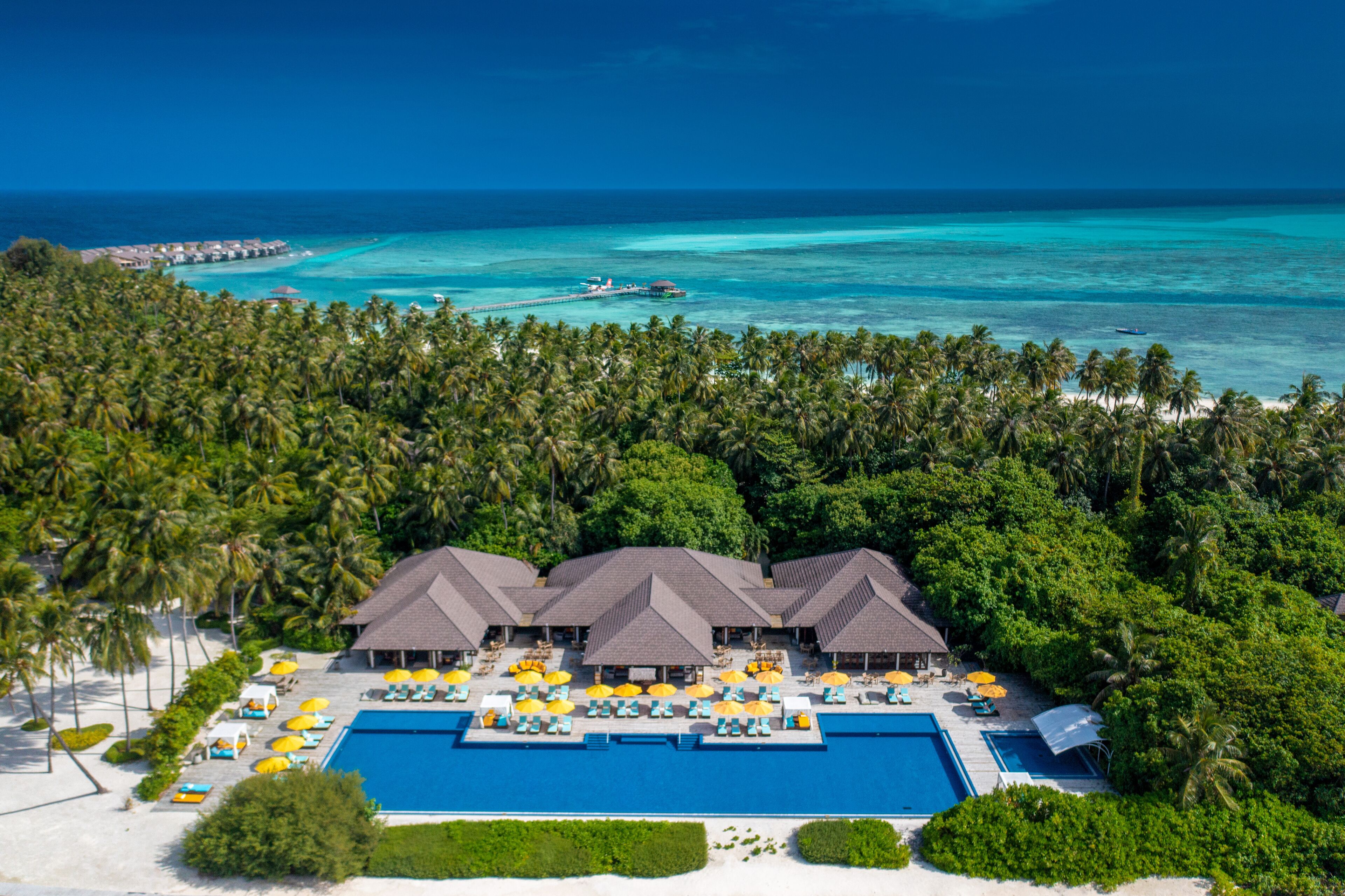 An aerial view of a resort with multiple overwater bungalows, a central pool, and a beach area.