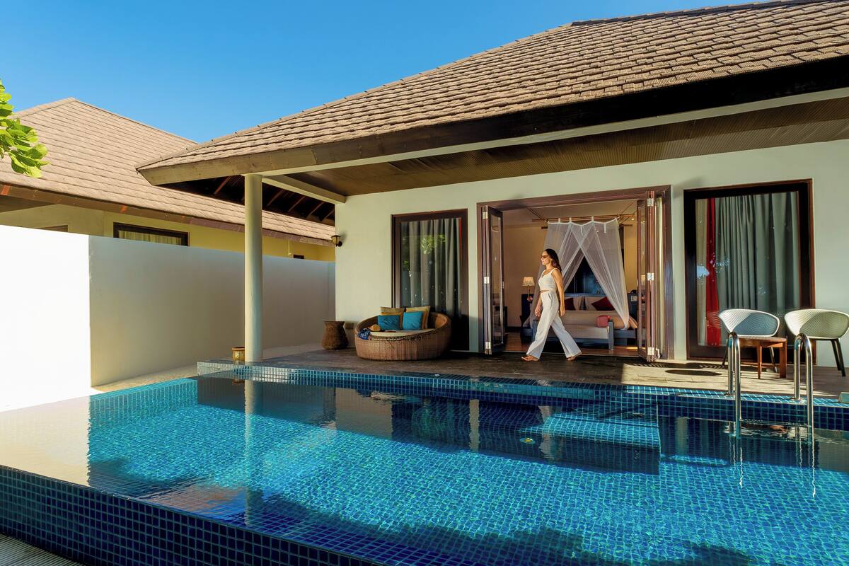 Ein Pool mit gefliestem Rand, ein überdachter Sitzbereich im Freien und ein Gebäude mit großen Fenstern und einem Ziegeldach.