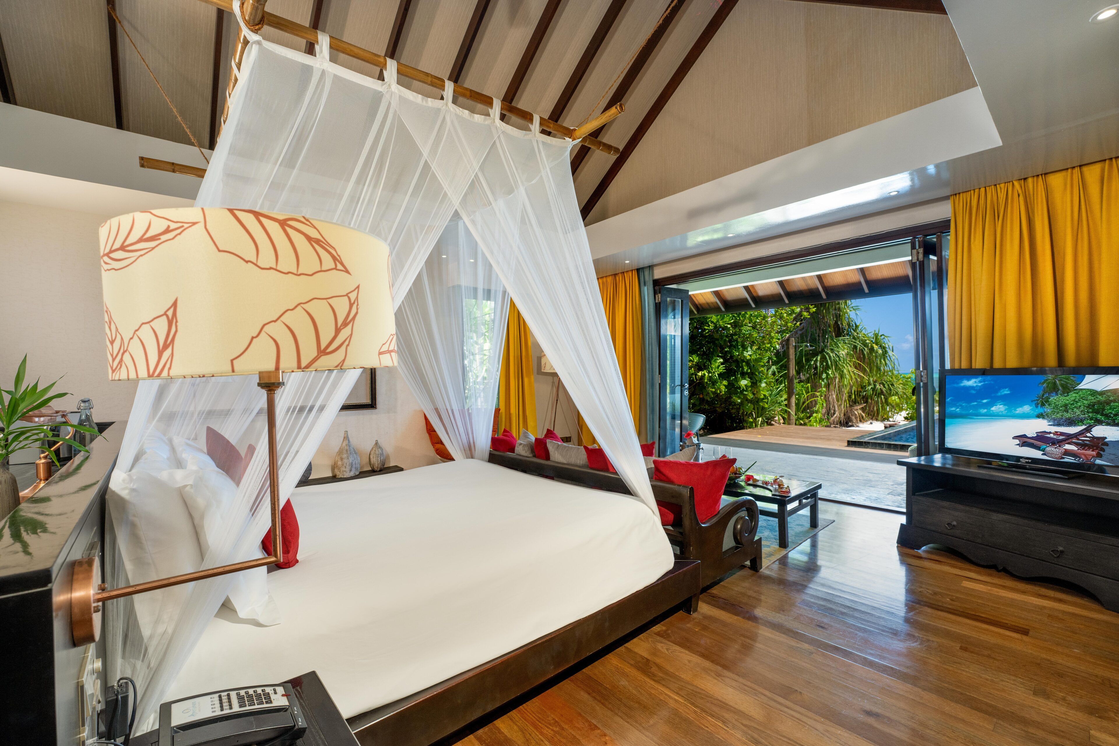 A modern bedroom with a bed, bedside table, TV, and a view of greenery outside.