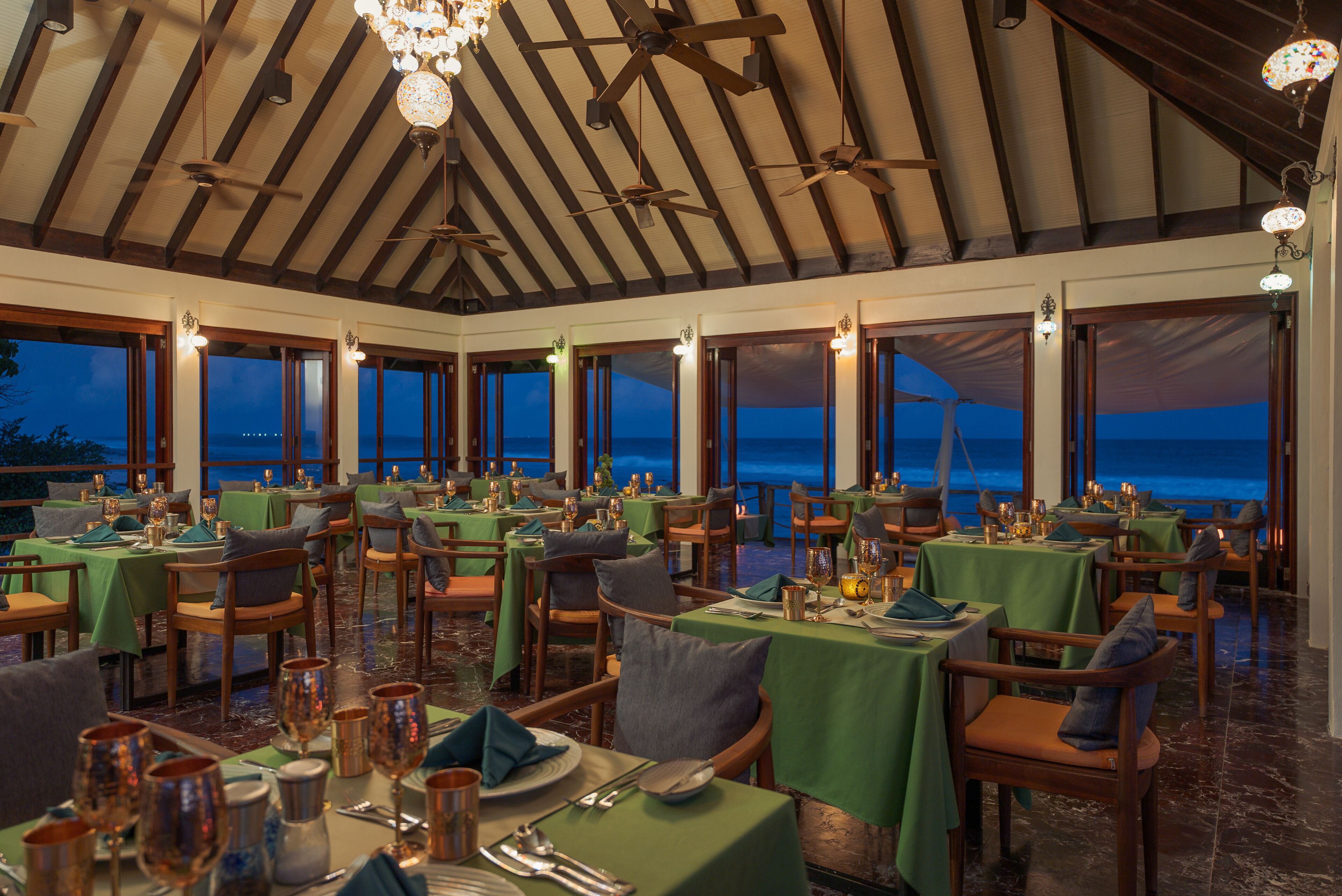 Un restaurante con varias mesas dispuestas para comer, amplios ventanales con vista al océano y un alto techo con vigas a la vista y ventiladores.