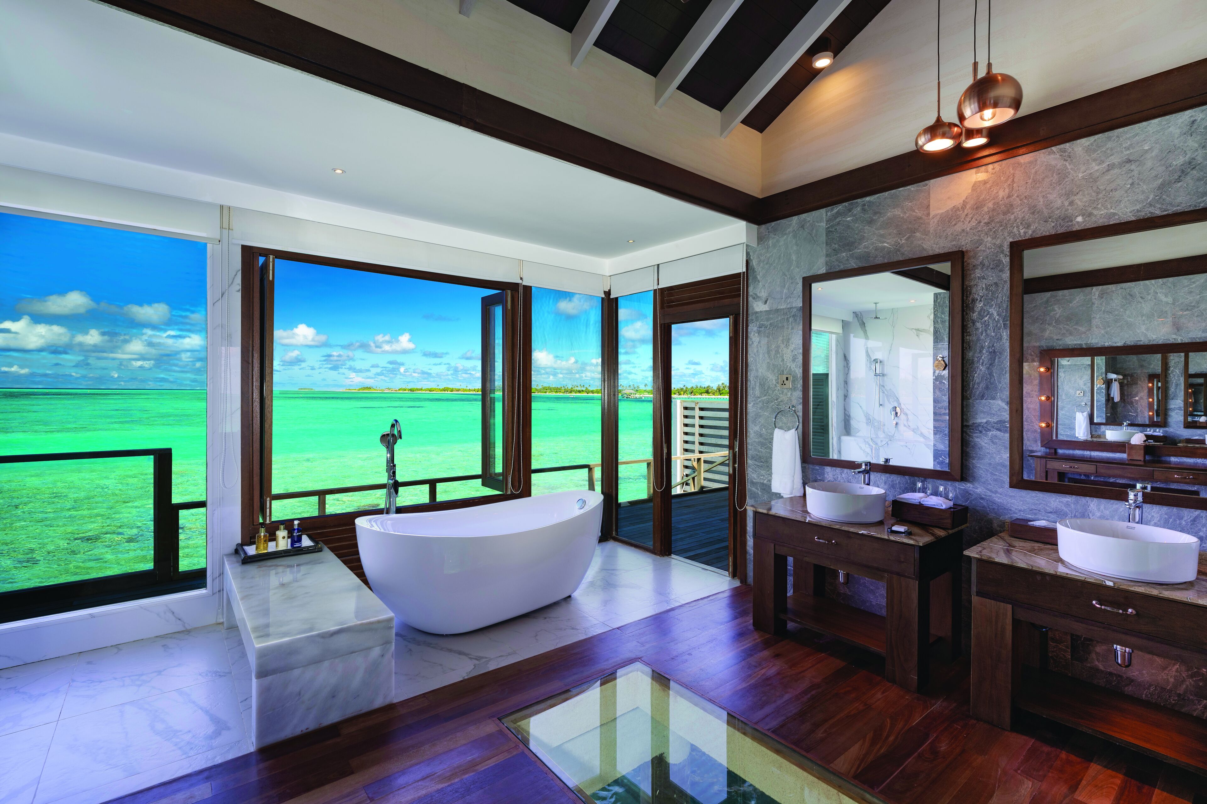 Un baño moderno con una bañera grande, dos lavamanos y un espejo amplio. Cuenta con vistas al océano a través de ventanas grandes.