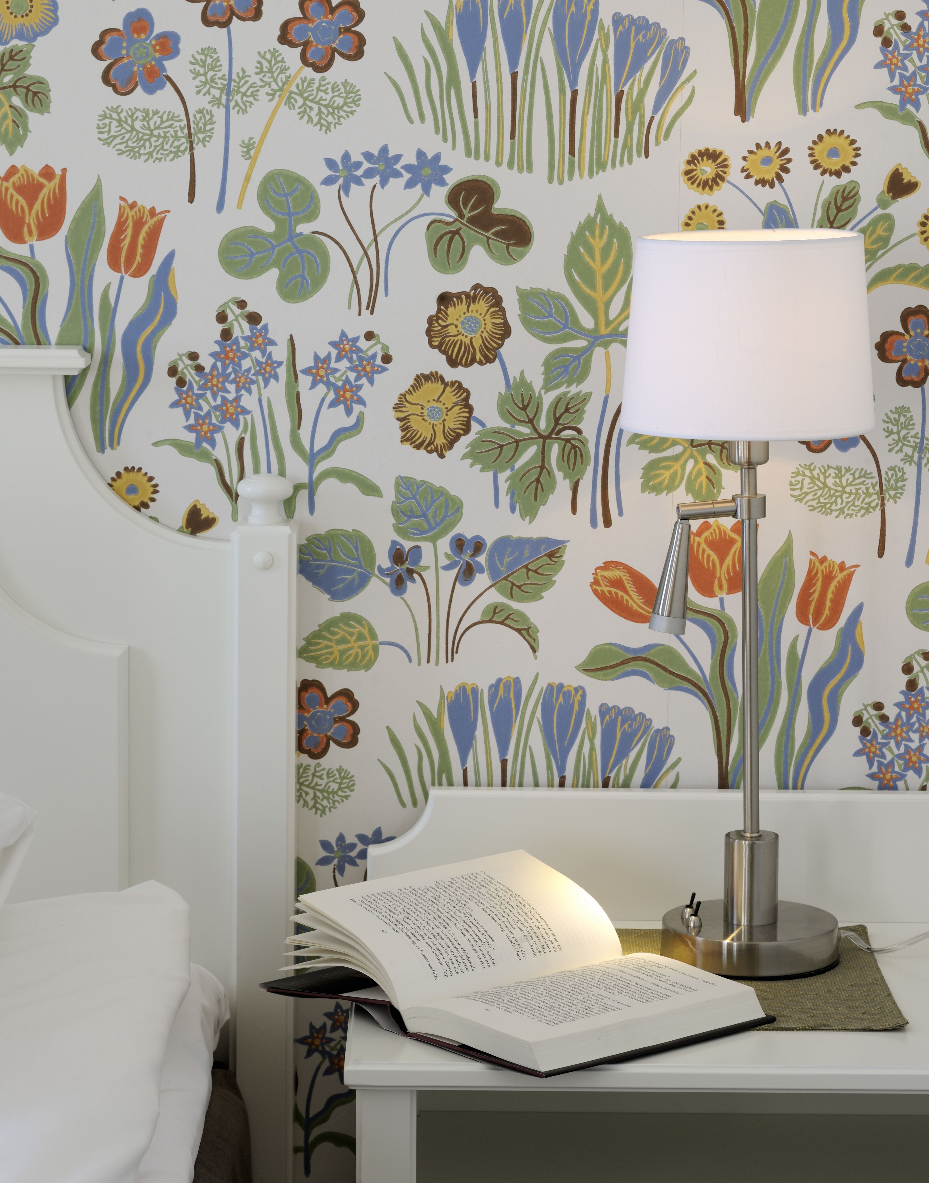 Ett rum med en vit säng, ett vitt skrivbord med en lampa och en vägg med blommig tapet.